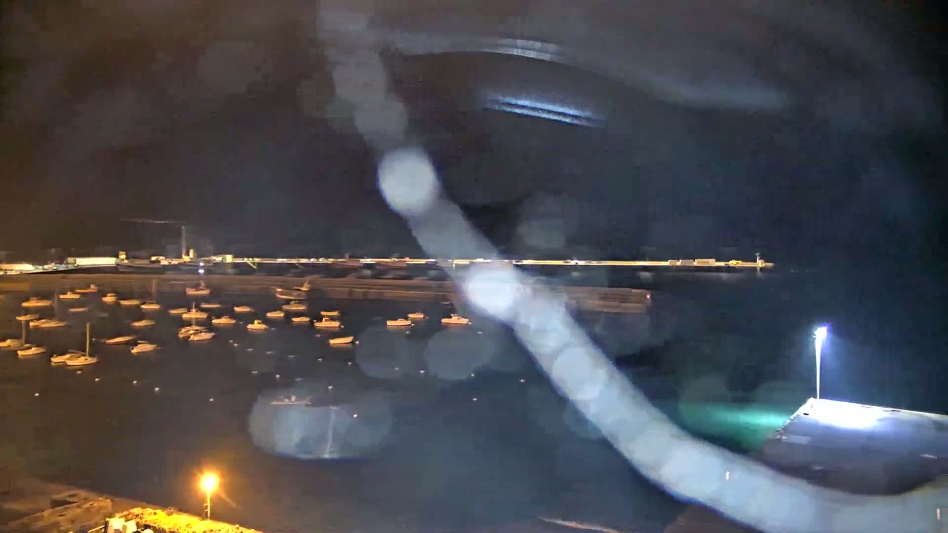 A nighttime view of a harbor filled with numerous small boats, viewed through rain-streaked glass.