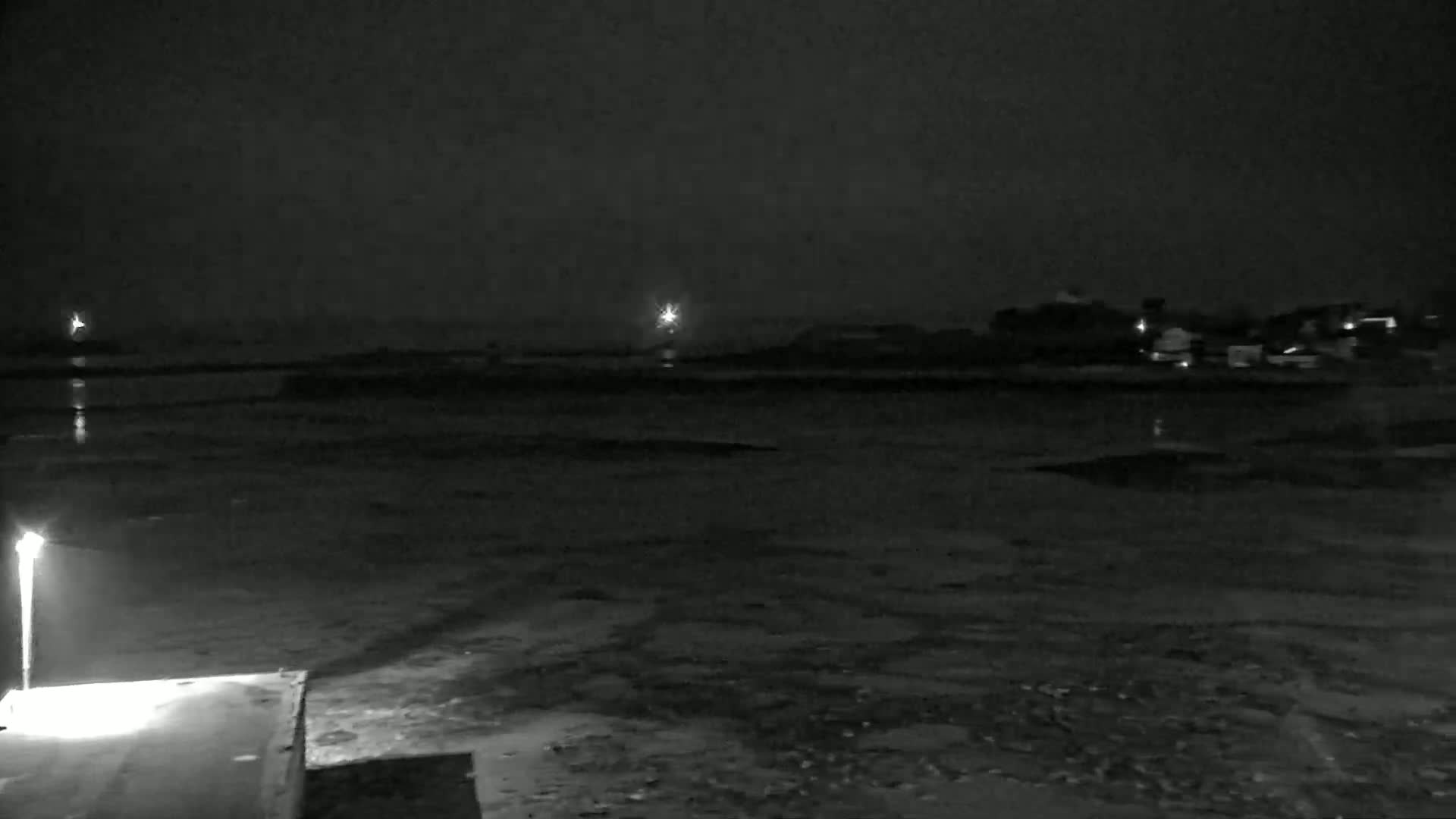 A nighttime, low-light view shows a dark body of water reflecting distant lights, with a darker shoreline and some structures visible in the background under a cloudy sky.