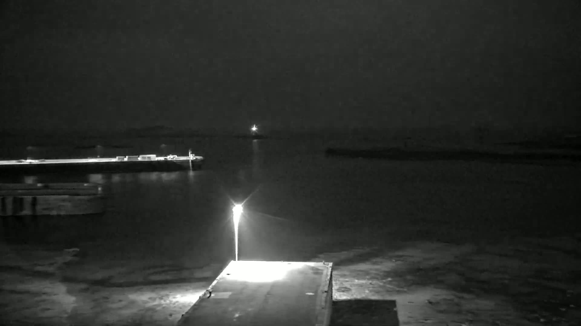 A nighttime, dark view of a harbor with a single lit lamppost on a dock, overlooking calm water and distant lights.