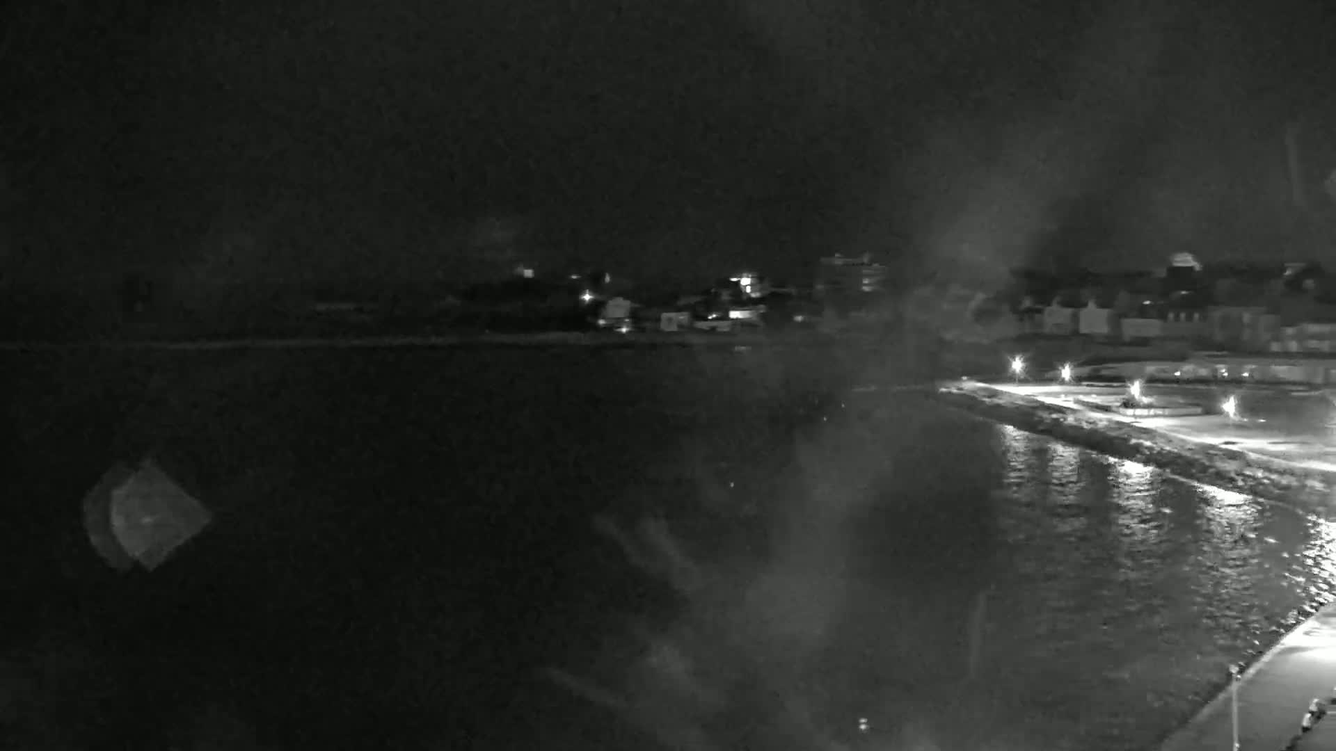 A nighttime, blurry view shows a dark body of water next to a dimly lit shoreline with buildings in the distance under a dark, possibly stormy sky.