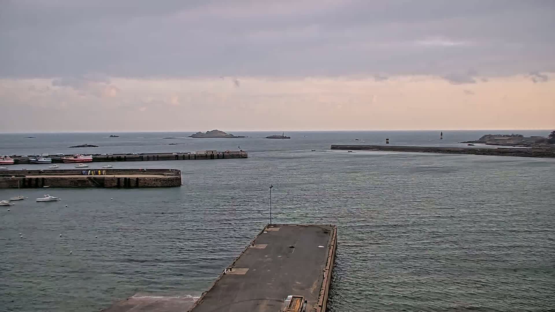 A harbor with several small boats, a long pier, and rocky islands in the distance under an overcast sky.