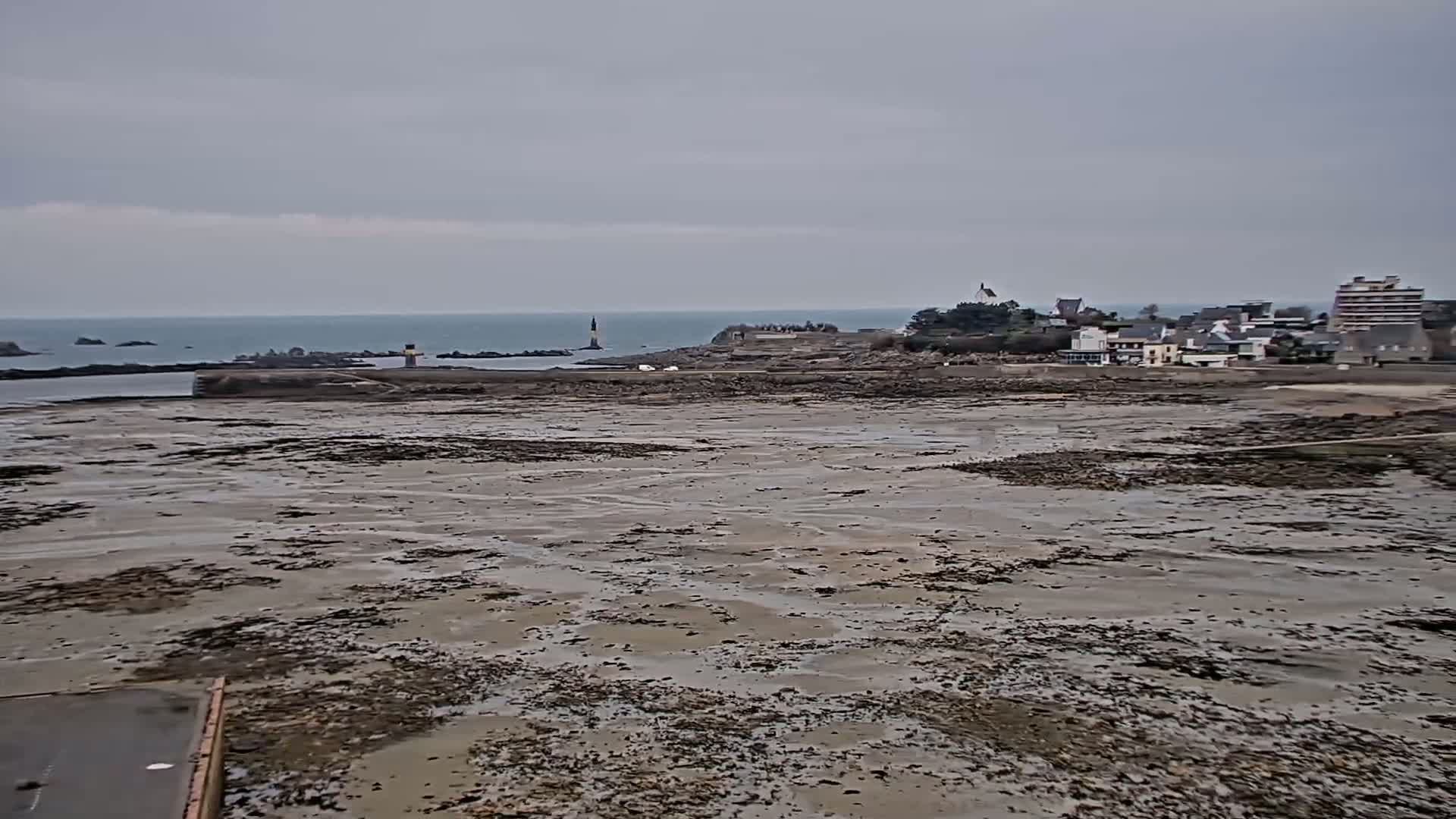 A wide shot of a coastal mudflat at low tide, with a small town and distant lighthouse visible under an overcast sky.