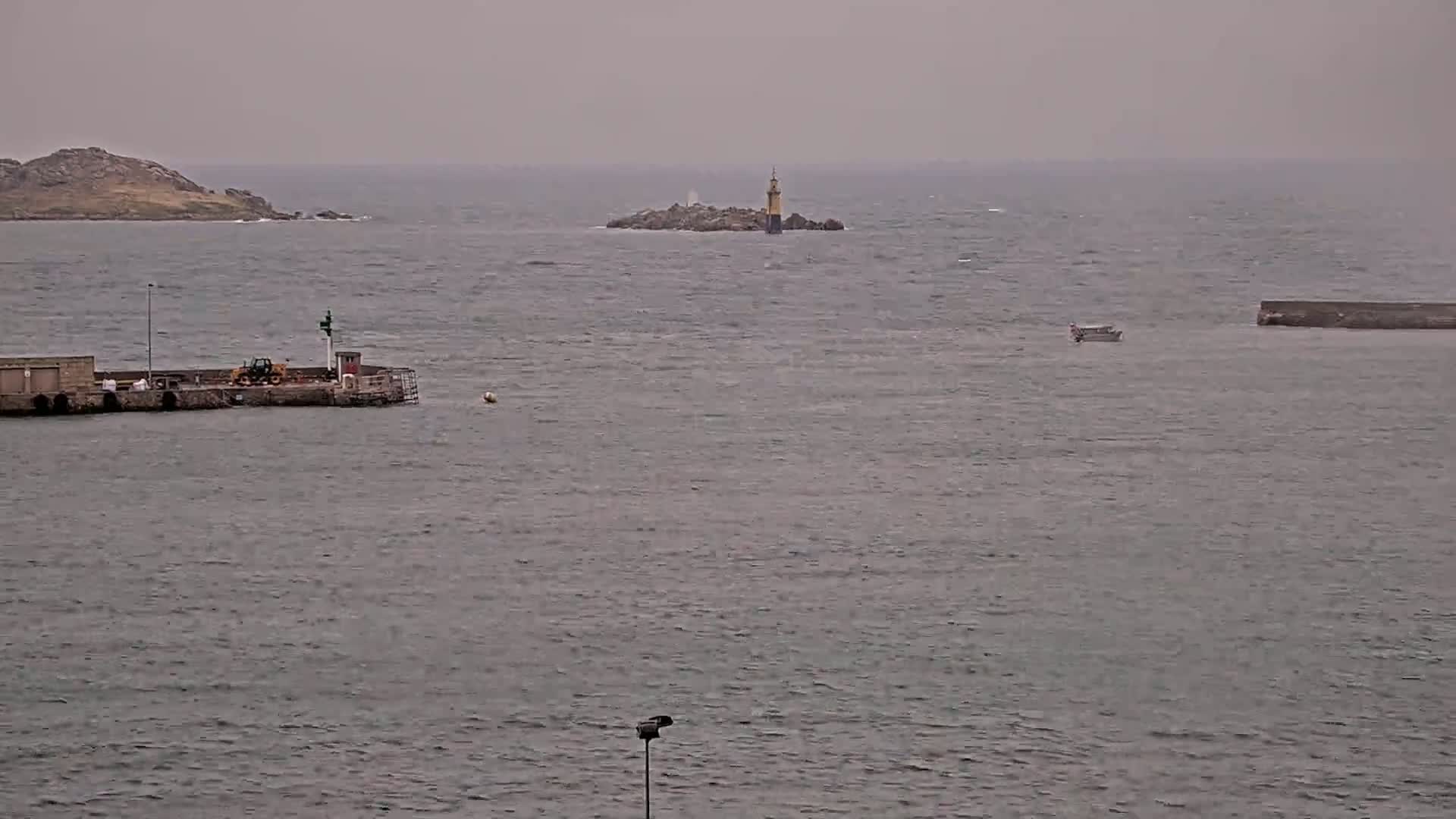 A small harbor with a lighthouse visible in the distance on a rocky island, under an overcast sky and slightly windy conditions.