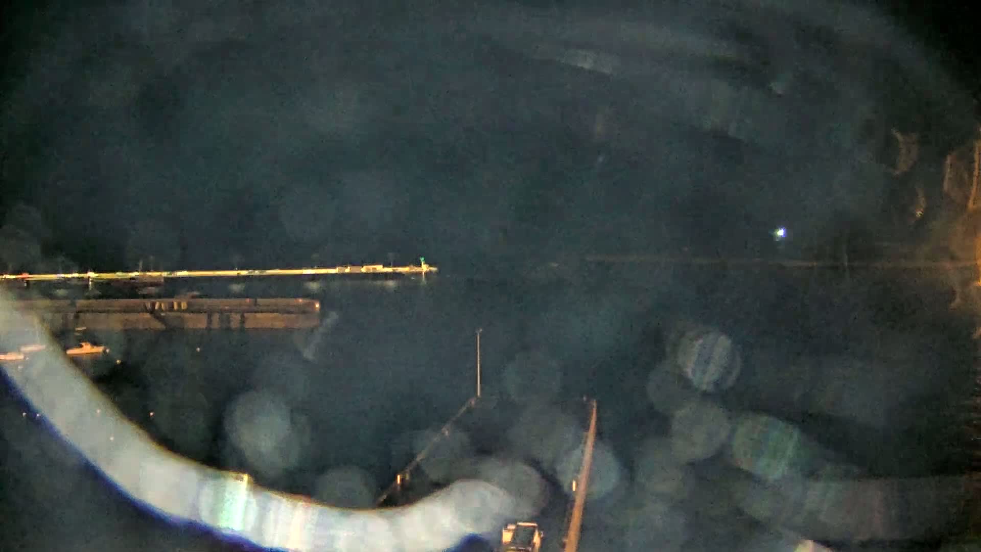 A nighttime view of a dark body of water with a lit-up pier in the distance, obscured by rain or fog.