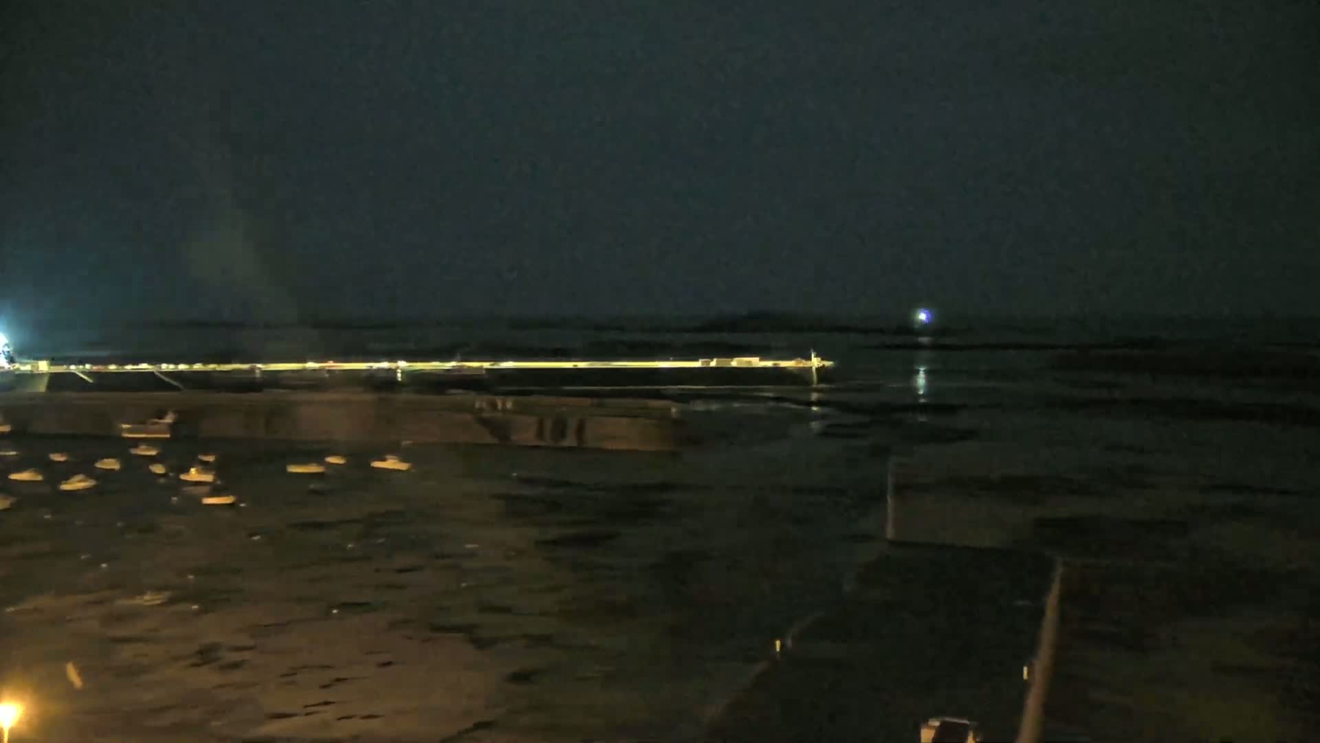 A nighttime view of a harbor with numerous small boats at rest, a distant illuminated structure, and a calm dark body of water under a dark sky.