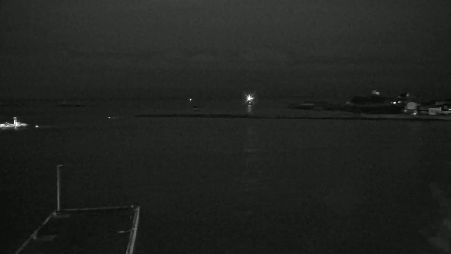 A dark, nighttime scene shows a body of water with faint lights on the horizon and a dark structure in the foreground under a cloudy sky.