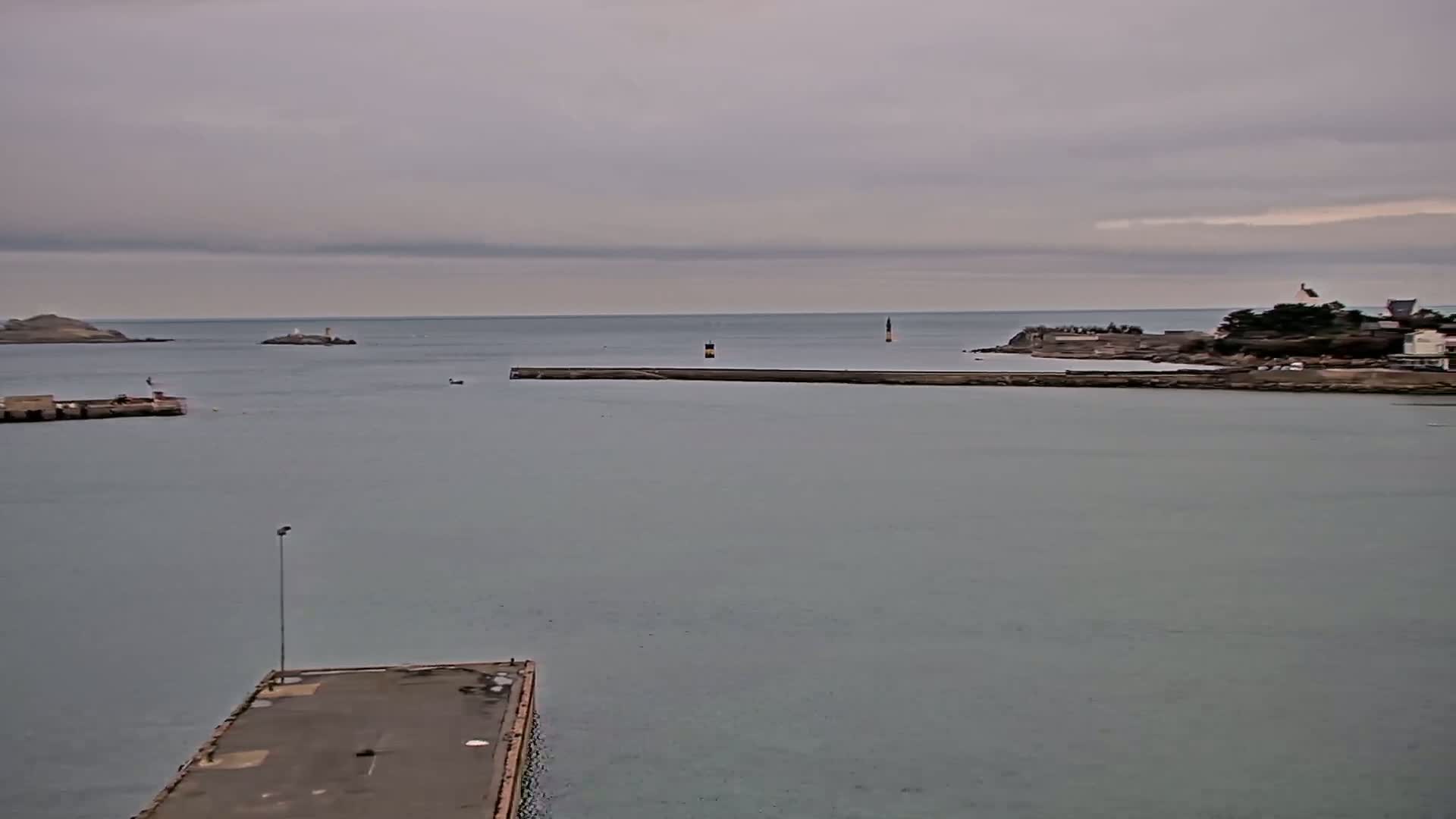 A calm, overcast day shows a harbor with a long jetty separating a small island with a lighthouse from a land-based harbor area.