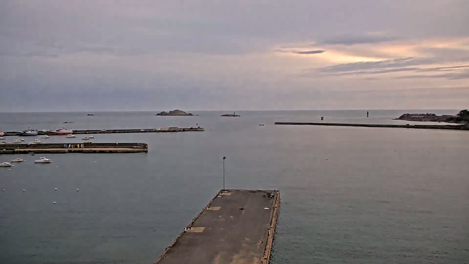A harbor with several boats docked, under a partly cloudy sky, shows a calm sea and small islands in the distance.