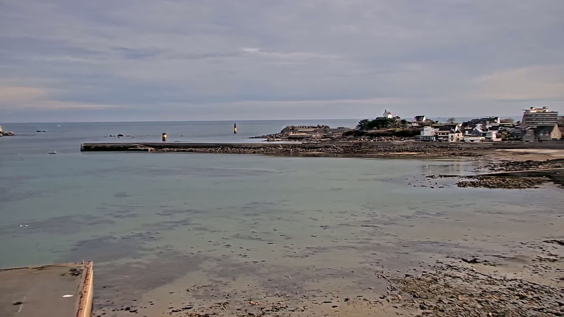 A calm, overcast day reveals a coastal town nestled beside a tranquil bay, partially exposed tidal flats, and a small harbor protected by a seawall.