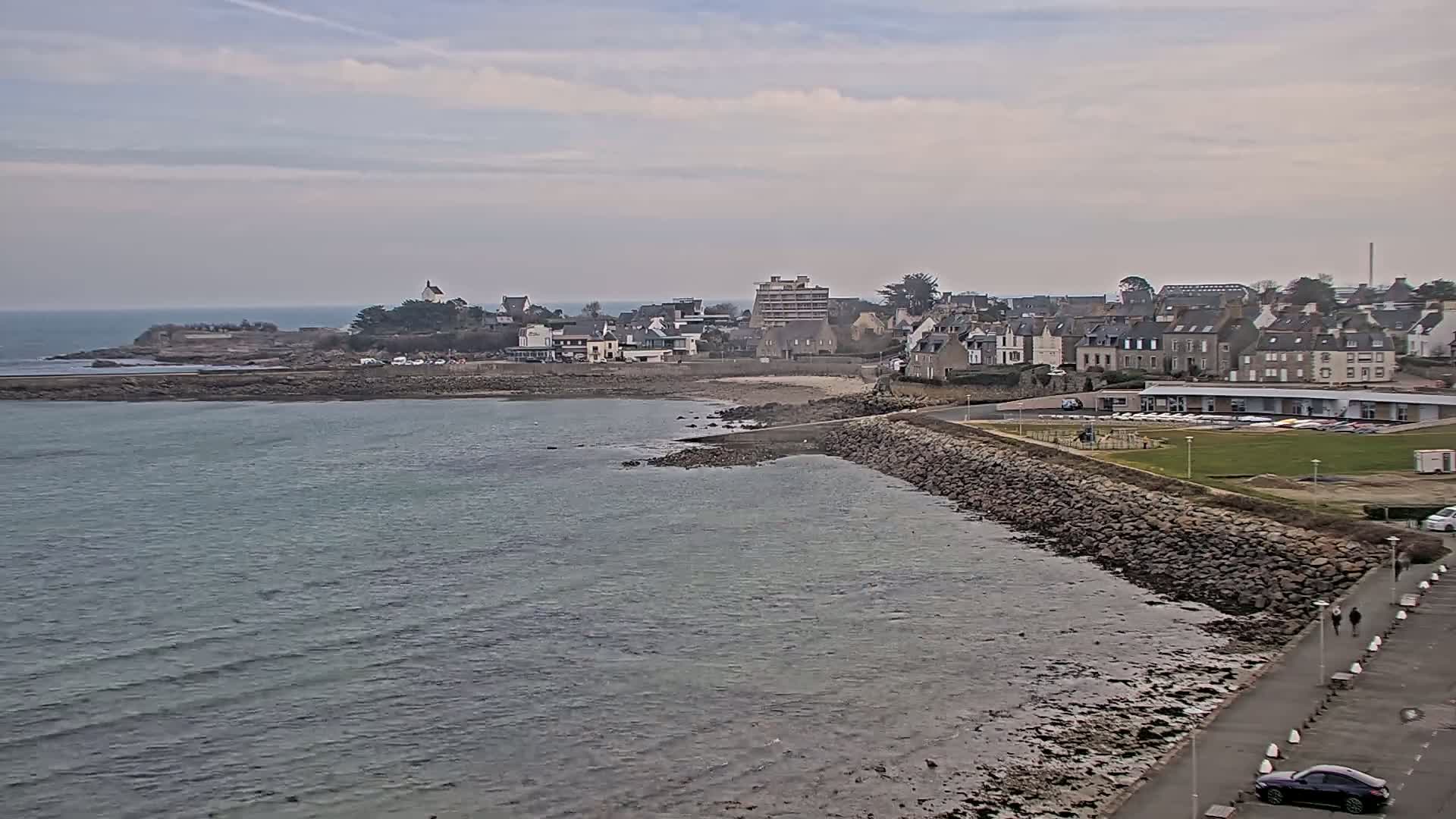A coastal town with buildings and a rocky shoreline is visible under a cloudy sky, with calm water extending to a calm sea in the background.