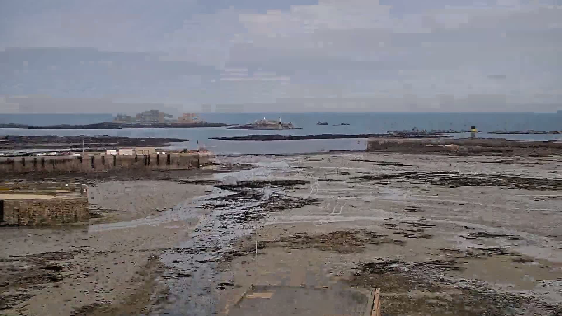 A low-tide coastal scene shows a stone harbor wall, exposed mudflats, and a distant island with a lighthouse under an overcast sky.