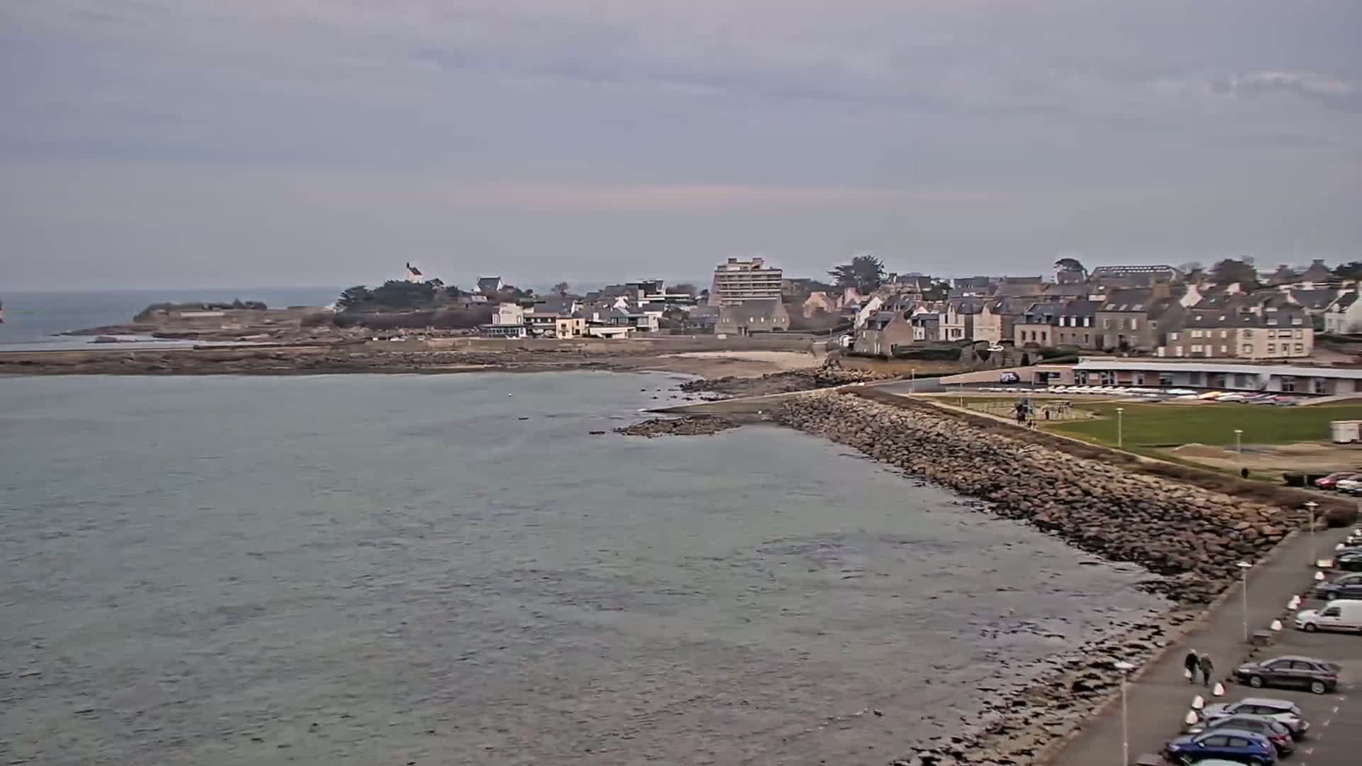 A coastal town with stone buildings sits on a rocky shoreline next to calm, gray water under an overcast sky.