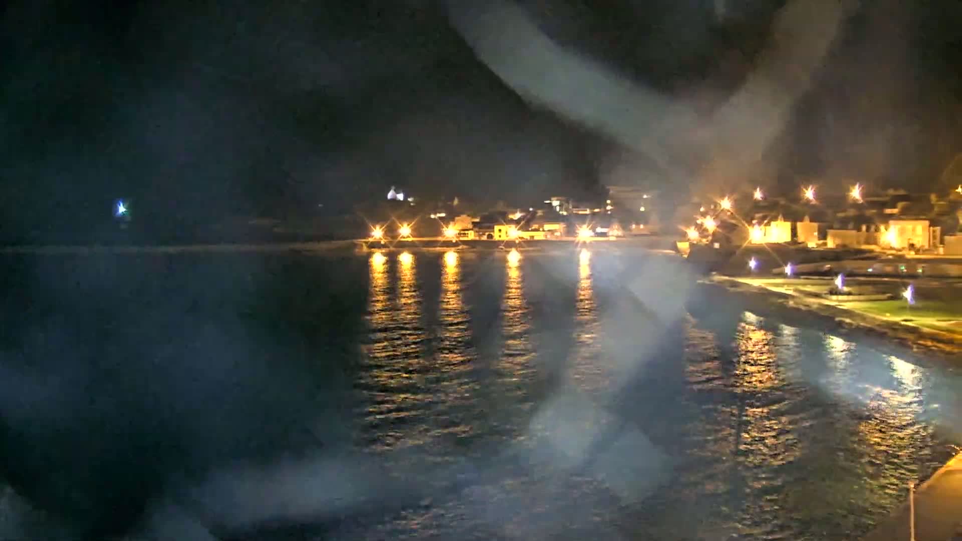 A nighttime view of a calm body of water reflecting the lights of a town on the opposite shore, slightly obscured by a hazy atmosphere.