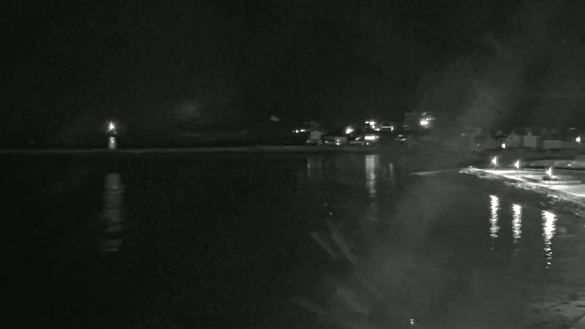 A nighttime view of a calm body of water reflecting lights from a shoreline with buildings in the distance under a dark, possibly foggy sky.