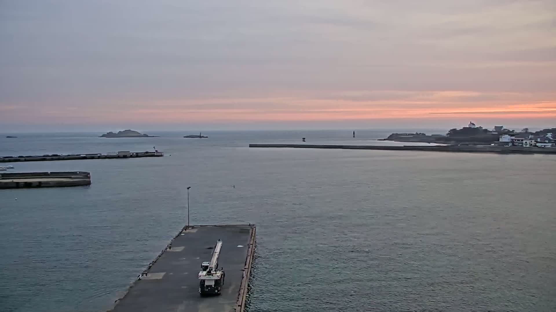 A harbor scene at sunset shows a crane on a dock, calm water, distant islands, and a small town on the shoreline.