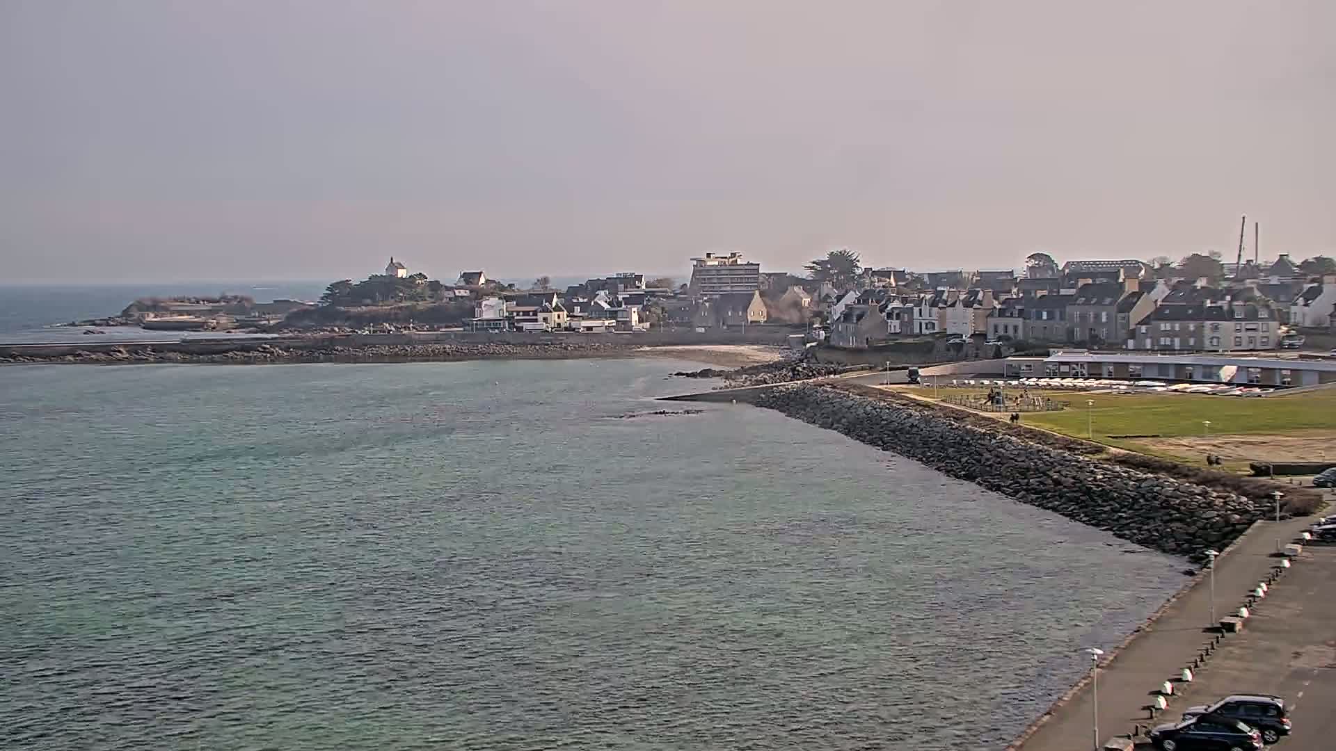 A coastal town with light-colored buildings and a rocky shoreline sits on a calm body of water under a hazy sky.