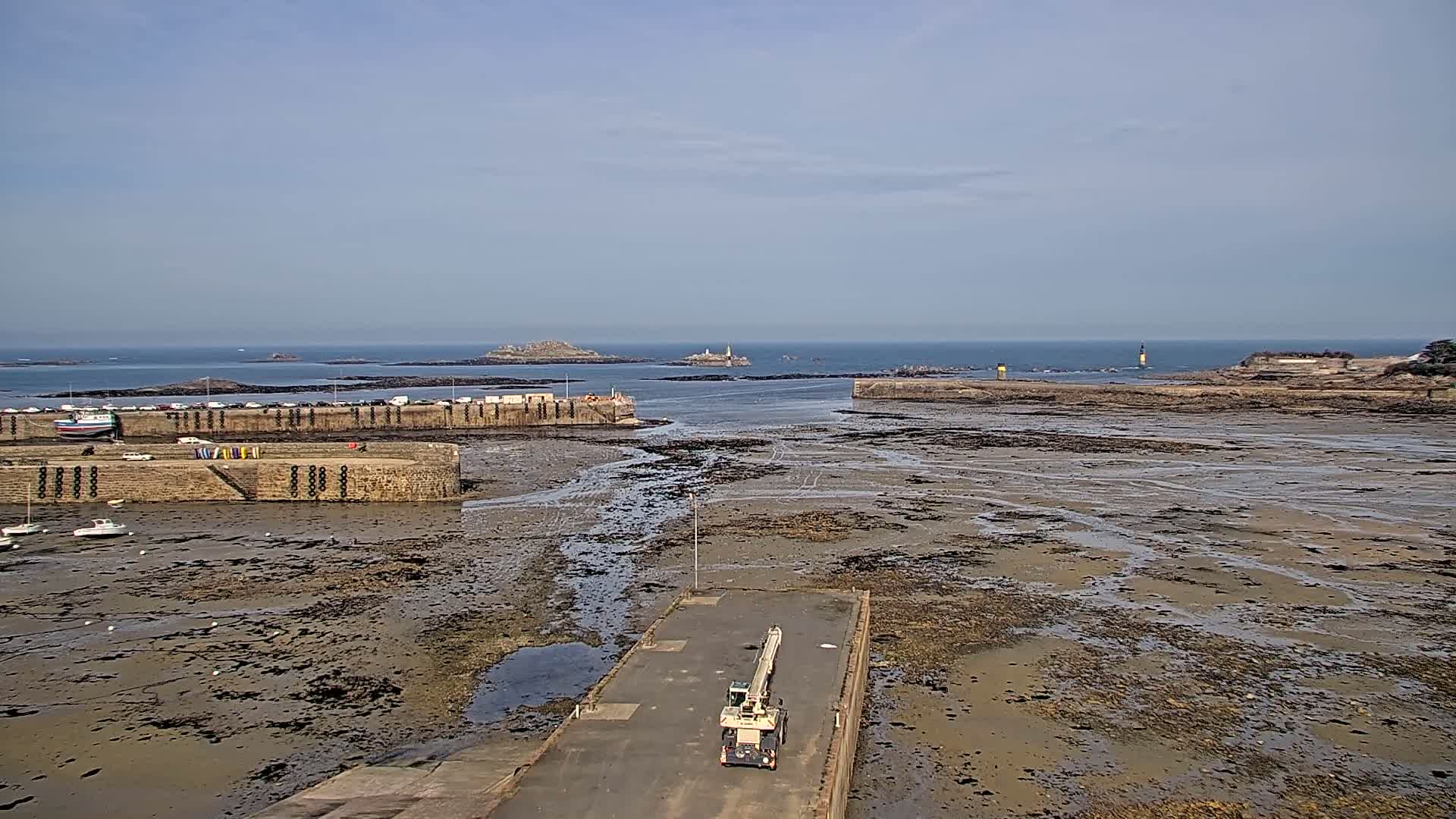 A harbor with a crane on a paved area, surrounded by exposed mudflats at low tide, overlooks islands and a calm ocean under a clear sky.