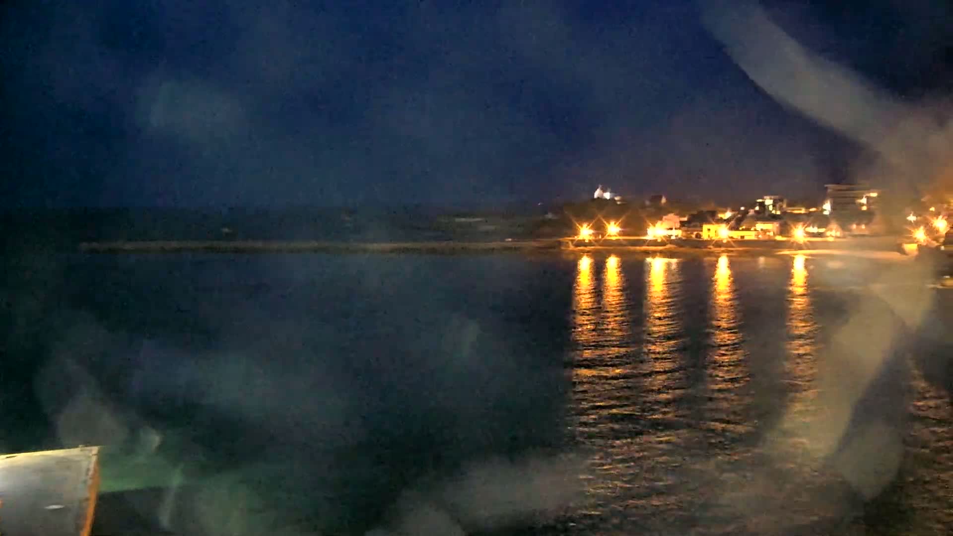 A nighttime view of a calm body of water reflecting the lights of a town on the shore under a dark sky.