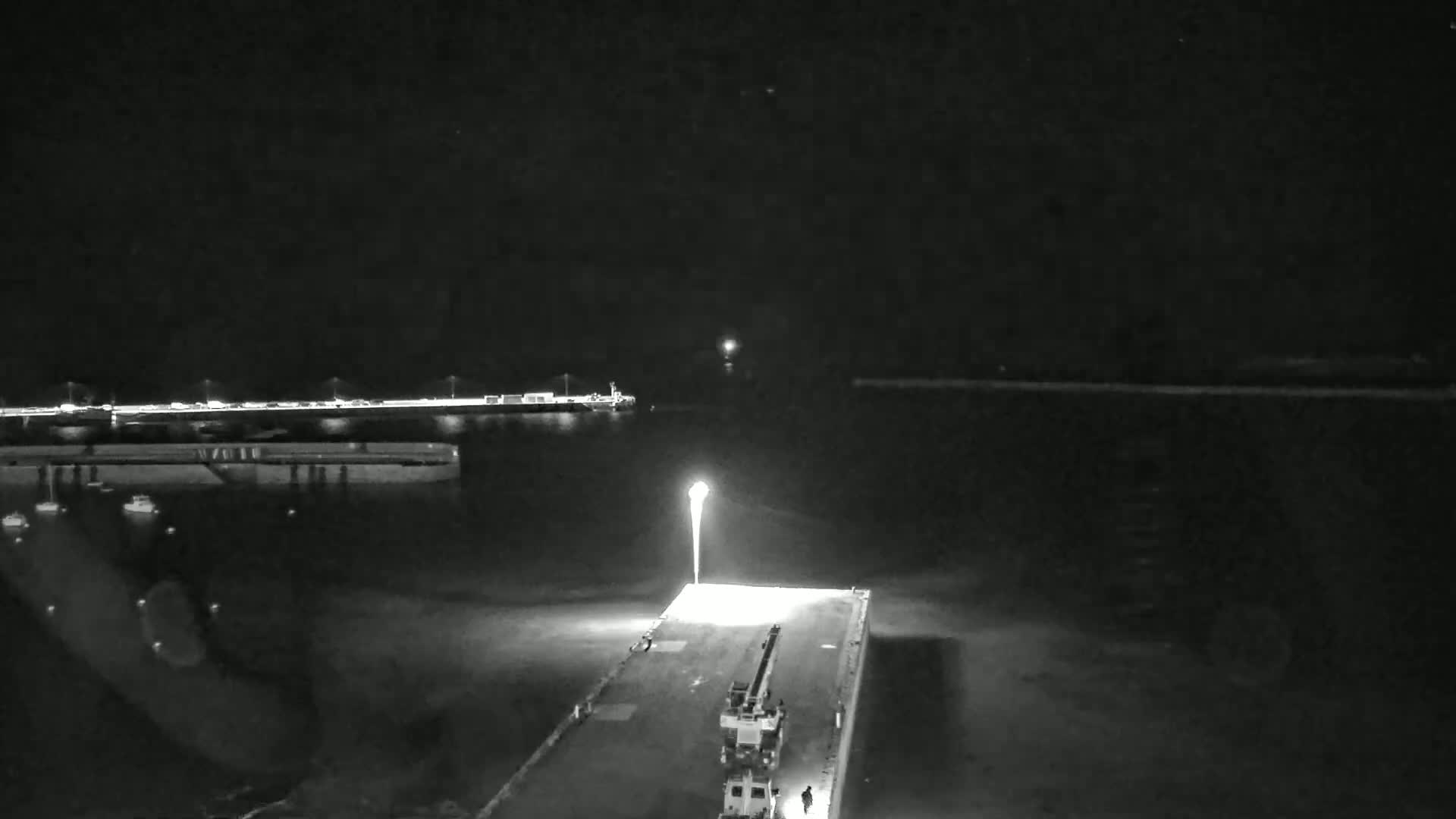 A nighttime, low-angle view shows a crane on a dock illuminated by a bright light, with calm water and a distant shoreline visible under a dark sky.
