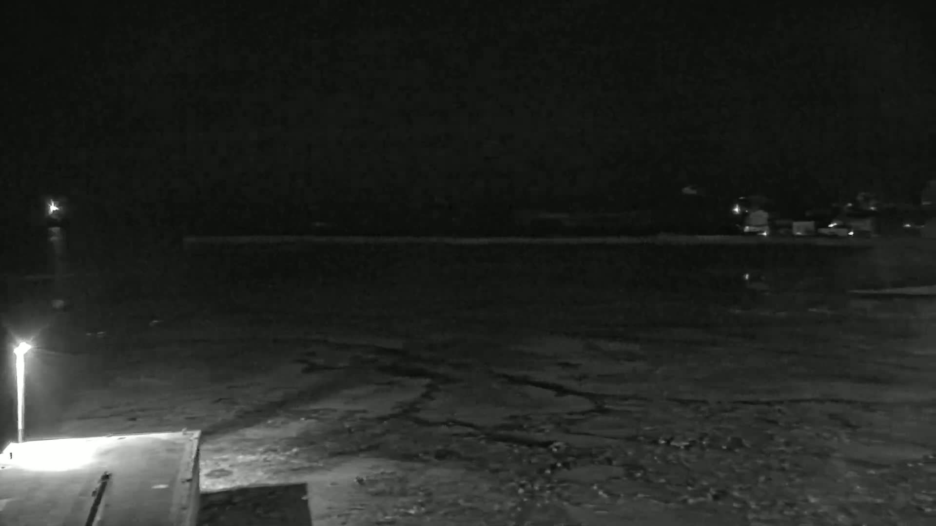 A nighttime, low-light image shows a dark body of water under a clear sky, with a dimly lit shoreline and a single lamppost illuminating a section of concrete.