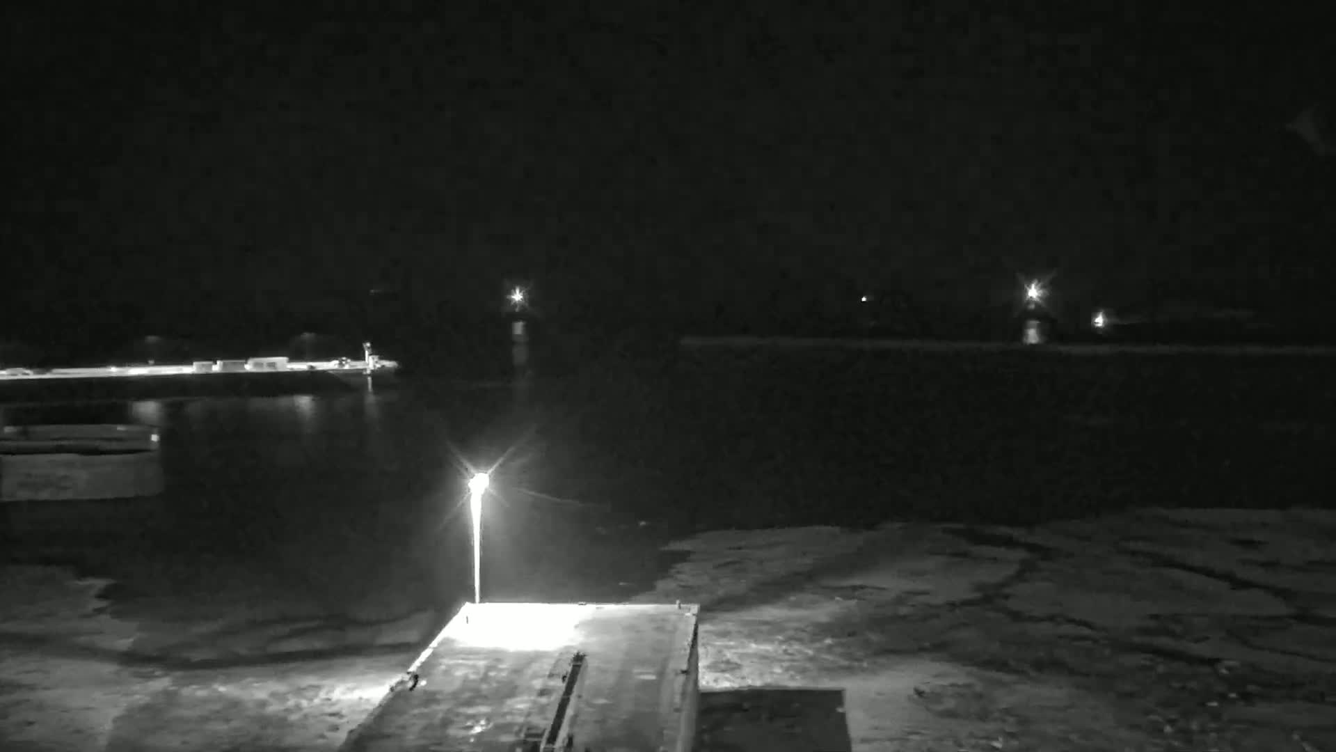 A nighttime view of a dark body of water, illuminated by a single lamppost on a dock, with distant lights visible across the water.