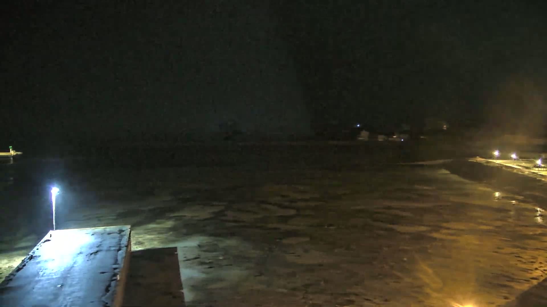 A nighttime, low-light view of a dark body of water, partially illuminated, with a pier and a few distant lights visible.