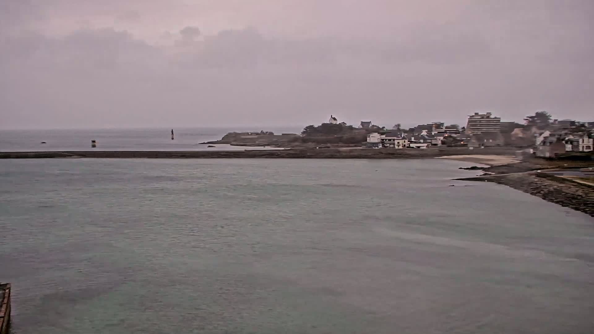 A coastal town under an overcast sky is partially visible across a calm body of water, with a breakwater extending into the sea.