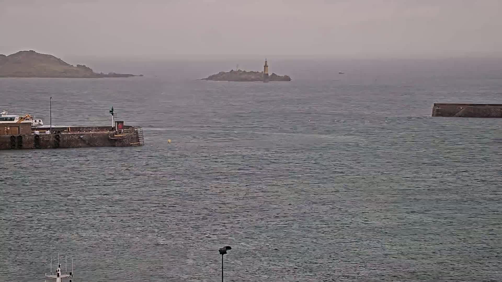A grey, overcast day shows a body of water with a small island featuring a lighthouse in the distance, a section of a harbor wall in the foreground, and a larger landmass further to the left.