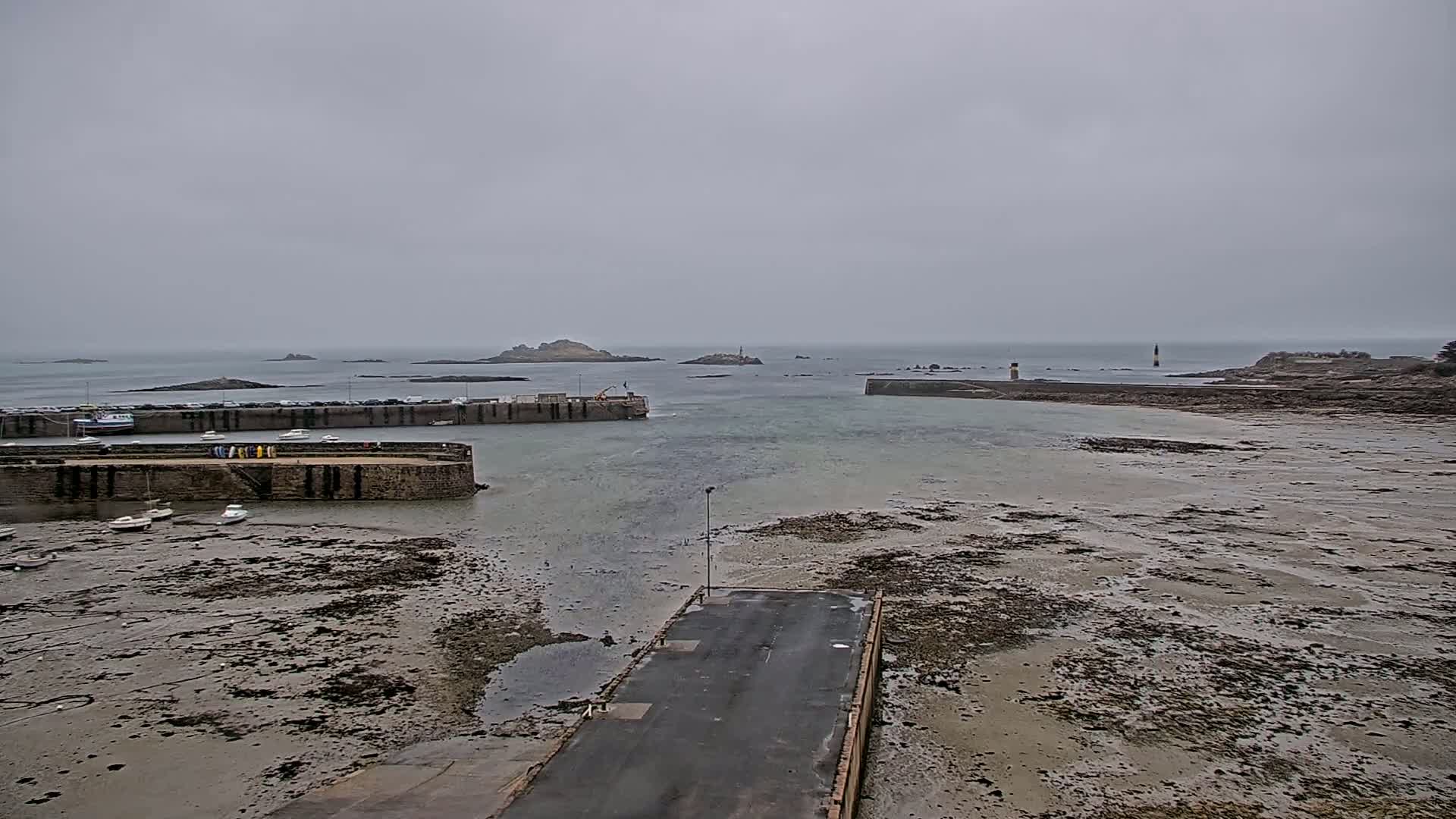 A harbor with several small boats is at low tide on an overcast day, with rocky islands visible in the distance.
