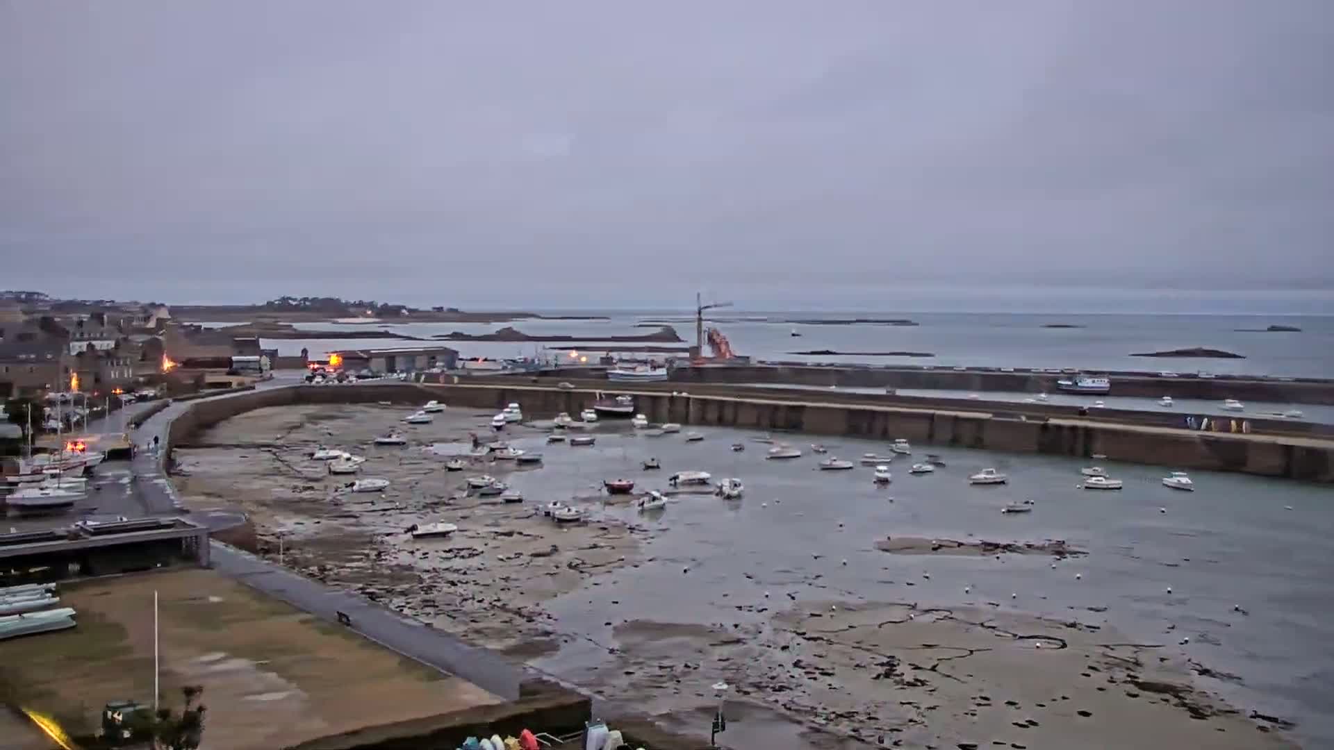 A harbor filled with numerous small boats sits exposed at low tide under an overcast sky, with a town and calm sea visible in the background.