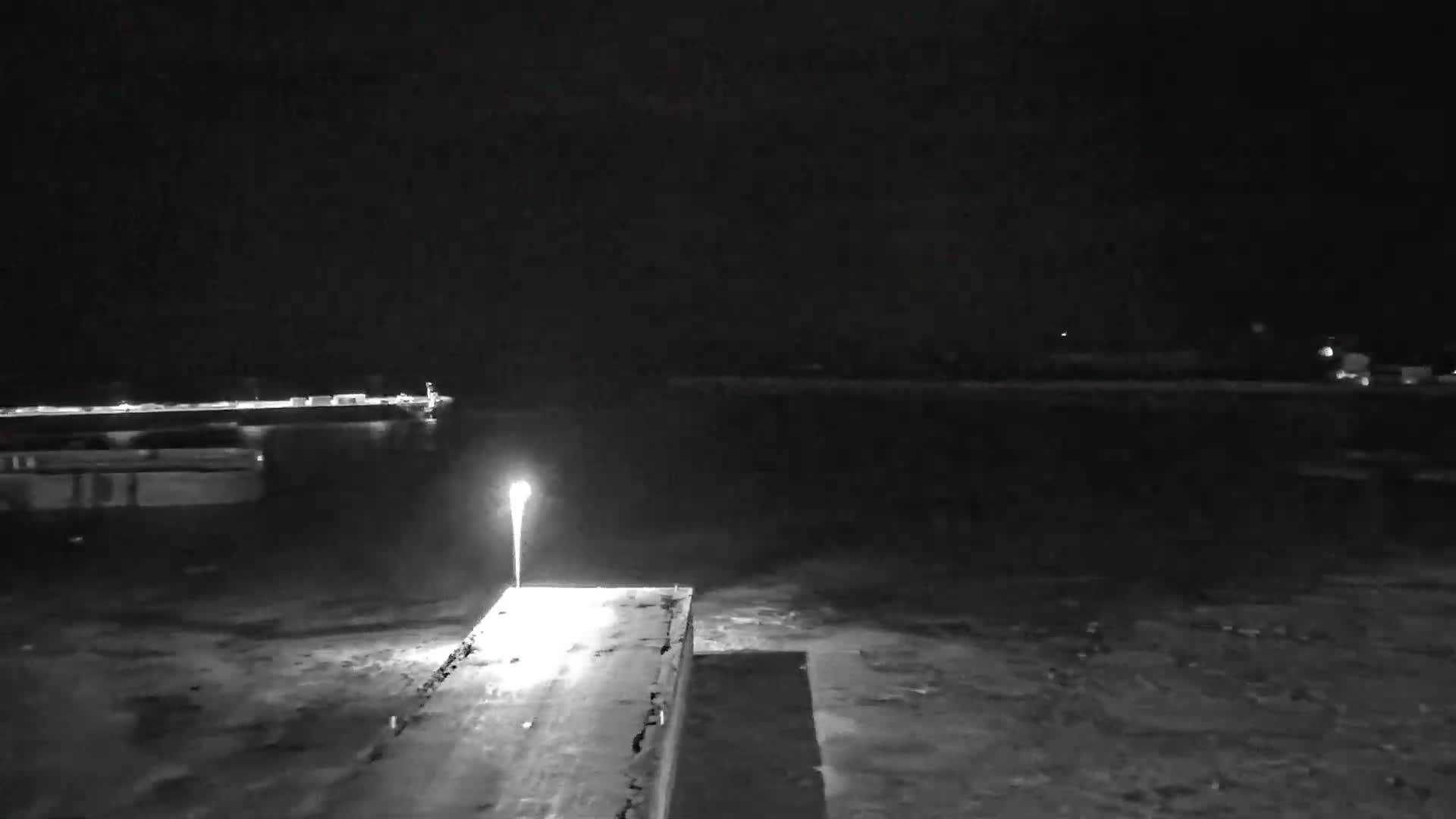 A nighttime view of a dark body of water, possibly a harbor, with a lit pier extending into the water, and faintly visible structures in the background.