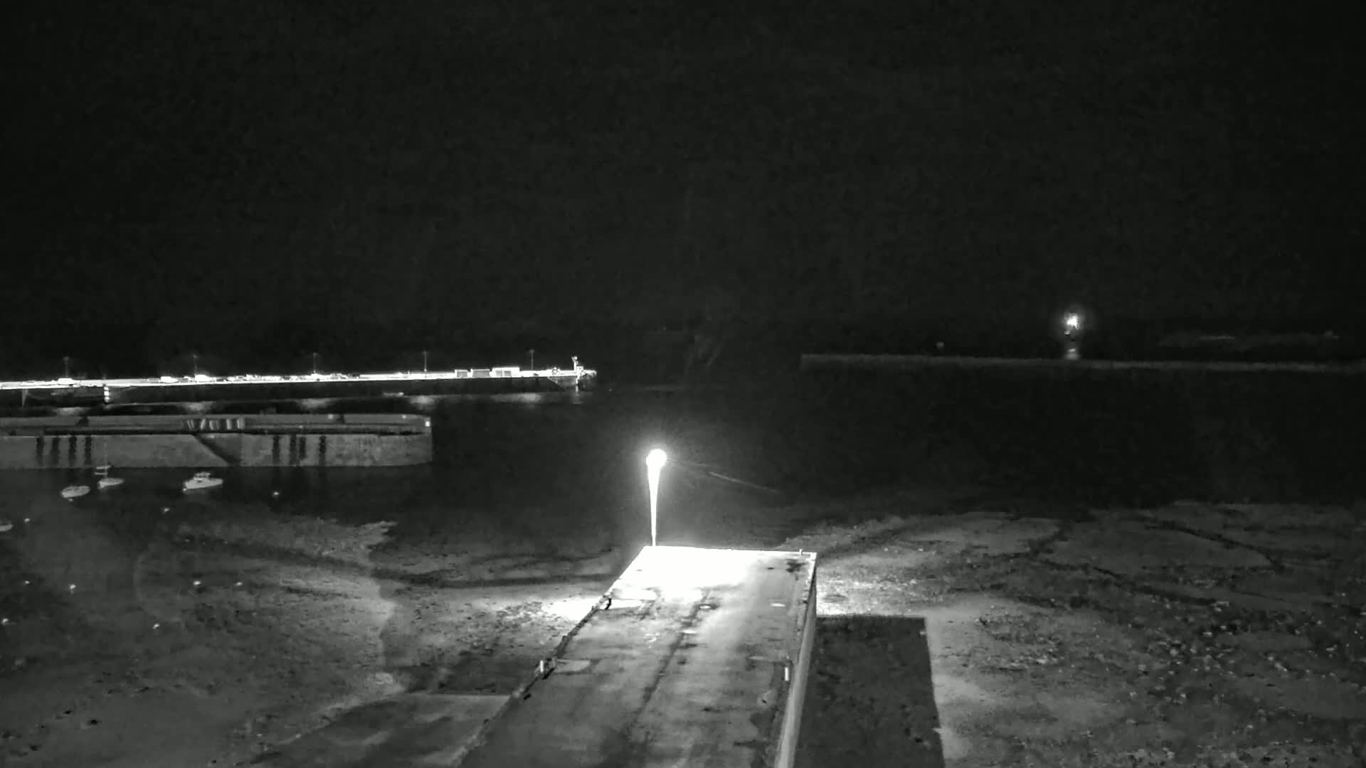 A nighttime view of a harbor with a single lit lamppost on a concrete platform, boats in the water, and a distant shoreline under a dark sky.