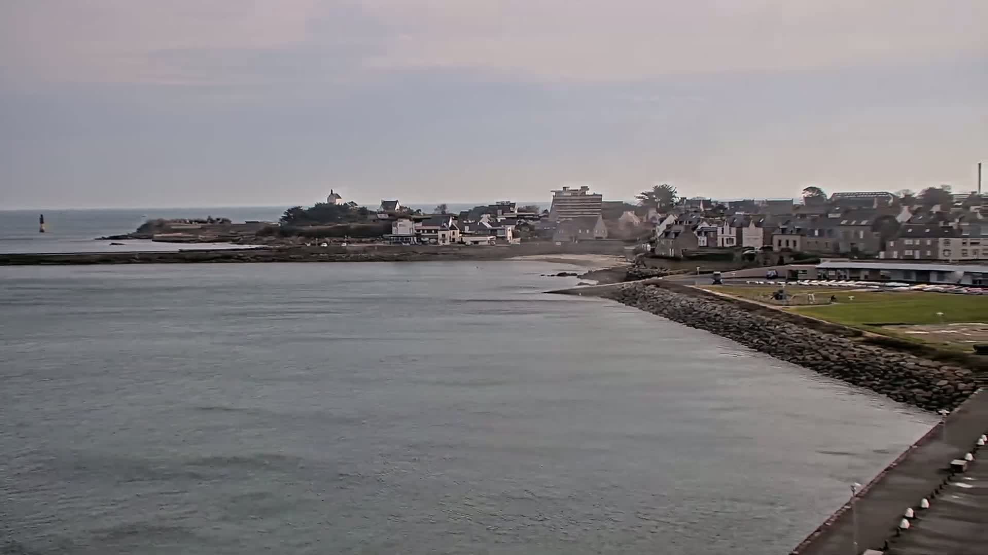 A coastal town with buildings and a small beach is visible across a calm body of water under an overcast sky.