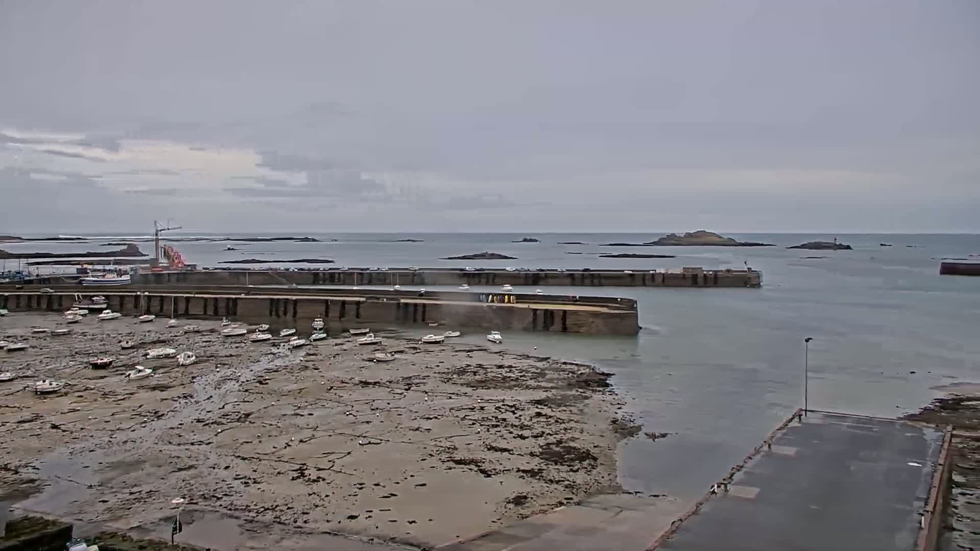 Under an overcast sky, a harbor filled with numerous small boats sits mostly exposed on a wide expanse of mudflats, with calm water surrounding a stone harbor wall and distant islands.