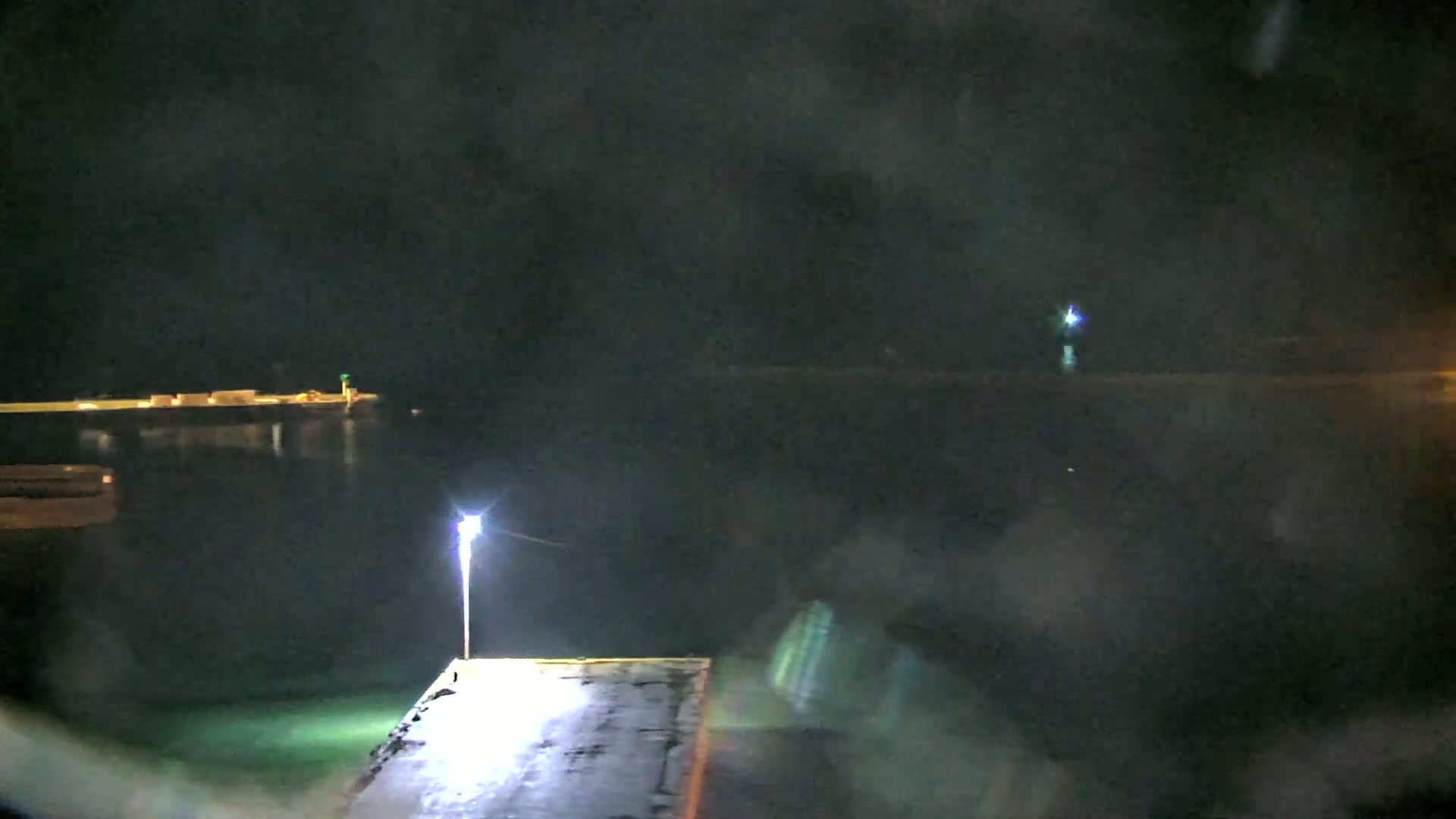 A nighttime view of a dark, calm body of water with a lit dock in the foreground and distant lights across the water under a slightly hazy sky.