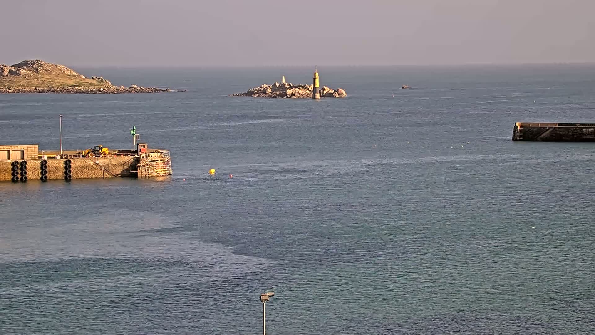 A harbor with a small, rocky island and lighthouse in the distance under a mostly clear sky.