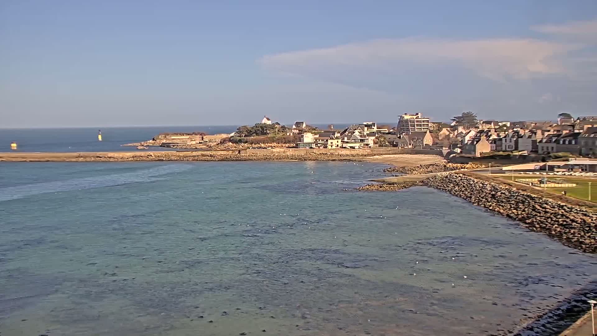 A coastal town with a small beach sits on a rocky peninsula under a mostly sunny sky, with calm, shallow, turquoise water surrounding it.