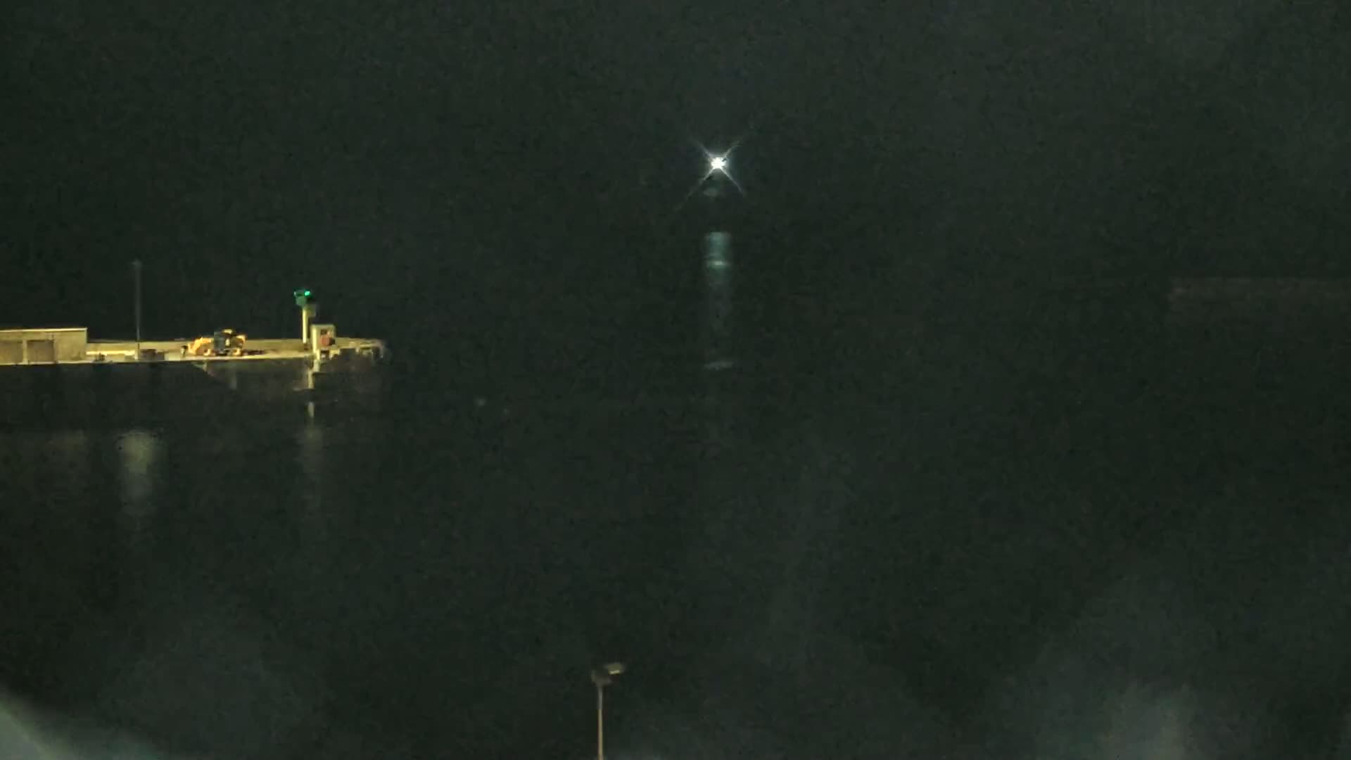A nighttime scene shows a dock with some machinery, a light on a pole, and a distant light source possibly a lighthouse, reflecting on dark water.