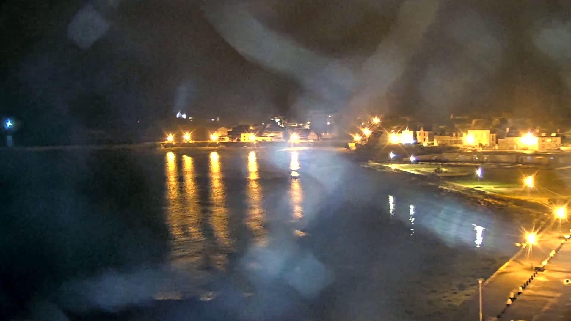 A nighttime view of a waterfront town, with its lights reflected on a calm body of water under a slightly hazy sky.