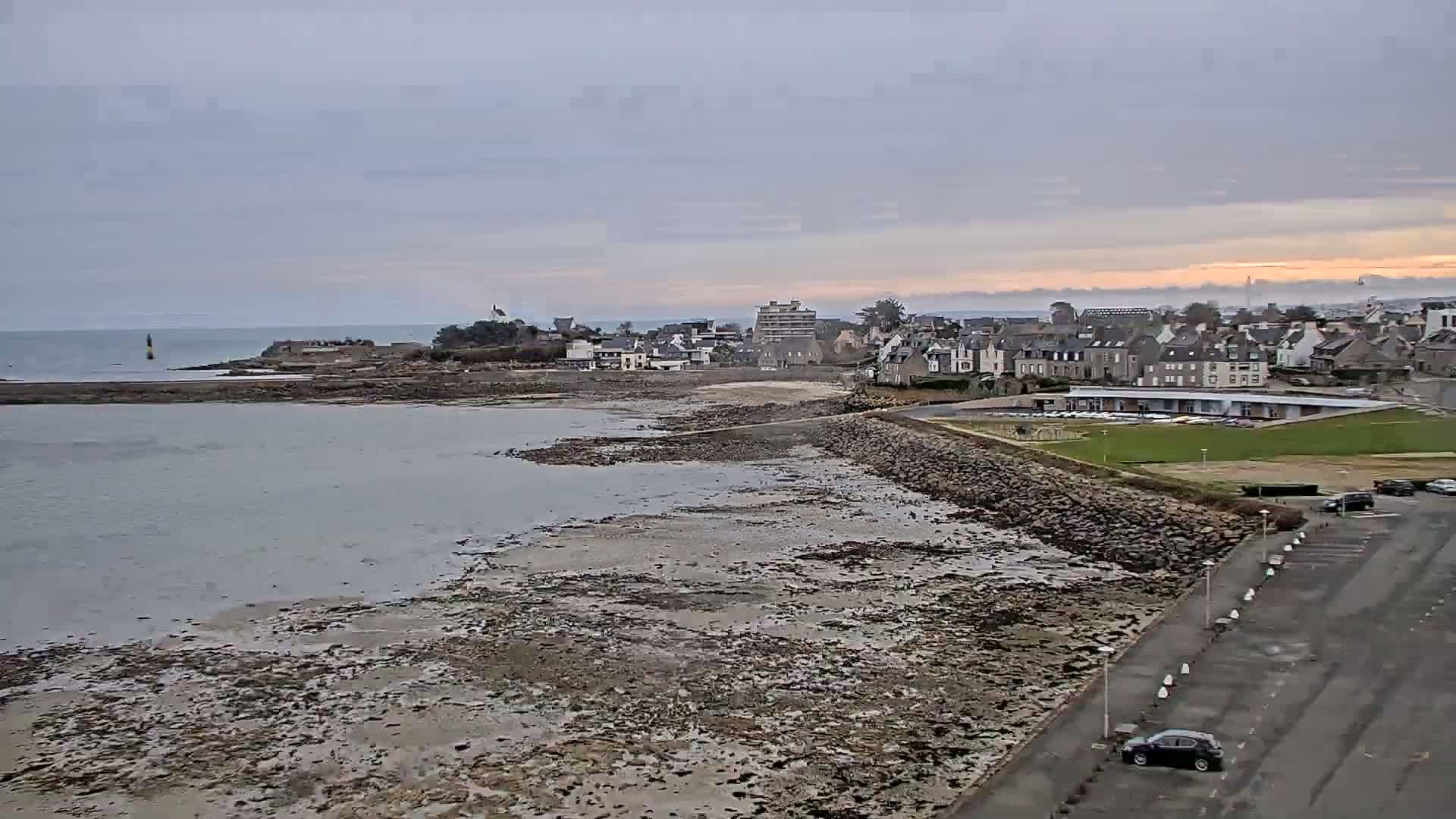 A coastal town is seen under a cloudy sky, with exposed mudflats and a calm body of water at low tide.