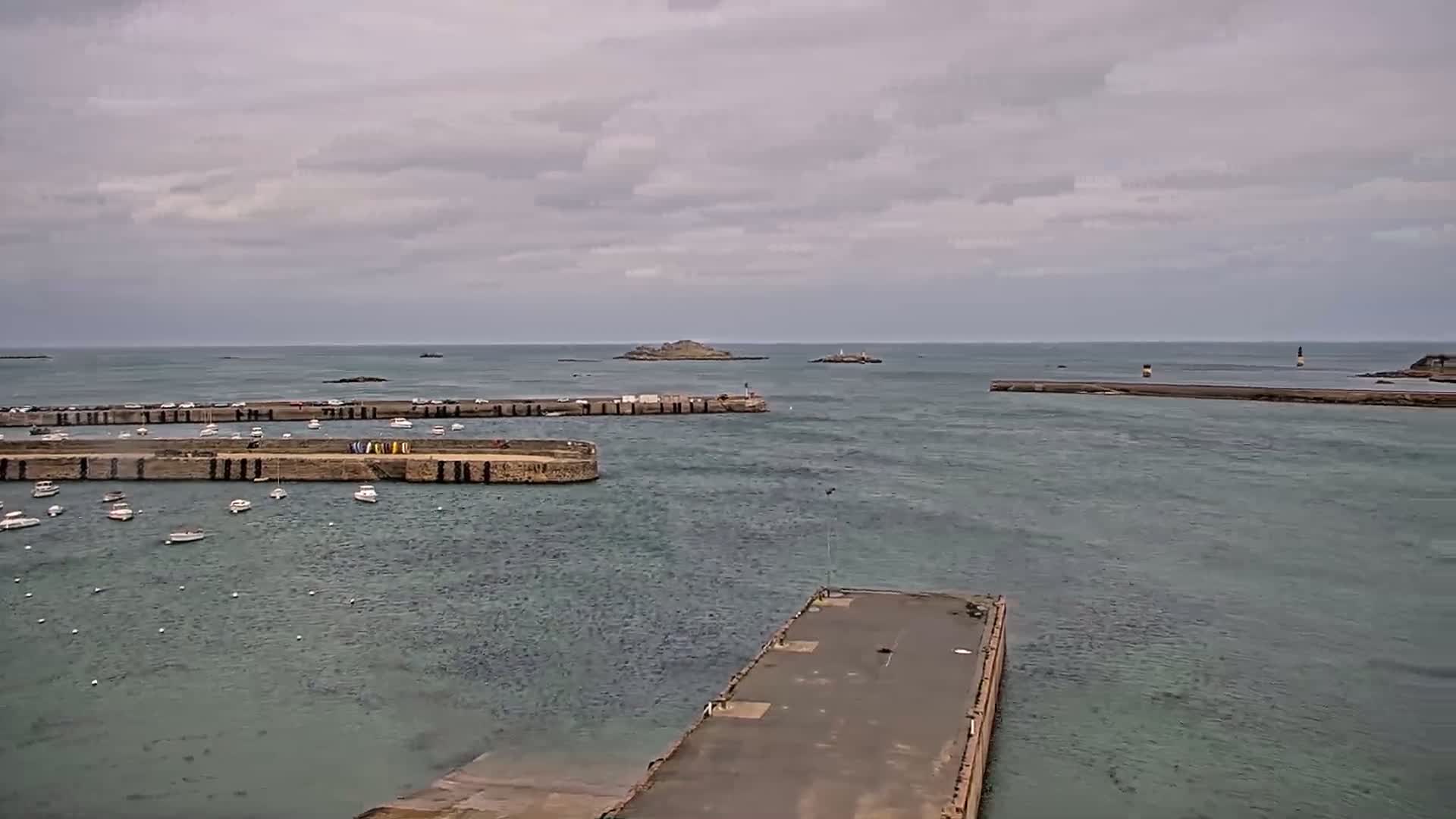 A harbor with numerous small boats is seen under an overcast sky, with a rocky island visible in the distance.