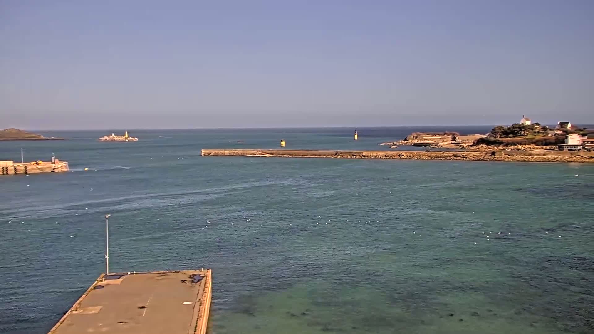 A tranquil harbor scene on a sunny day, with calm teal water, a long breakwater, small islands with lighthouses in the distance, and a small landmass with buildings in the background.