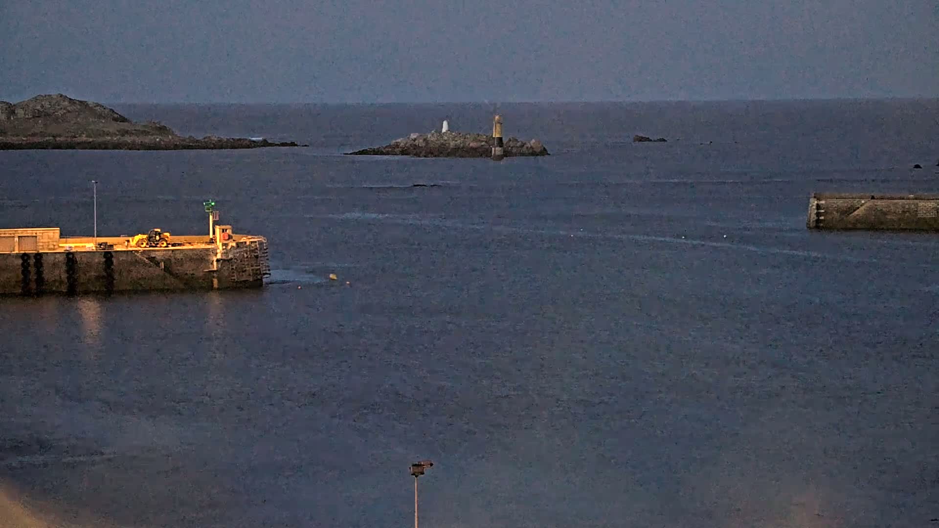 A twilight view of a harbor with a small island featuring two lighthouses in the distance, a piece of harbor wall to the left with a yellow vehicle, and another piece of harbor wall to the right.