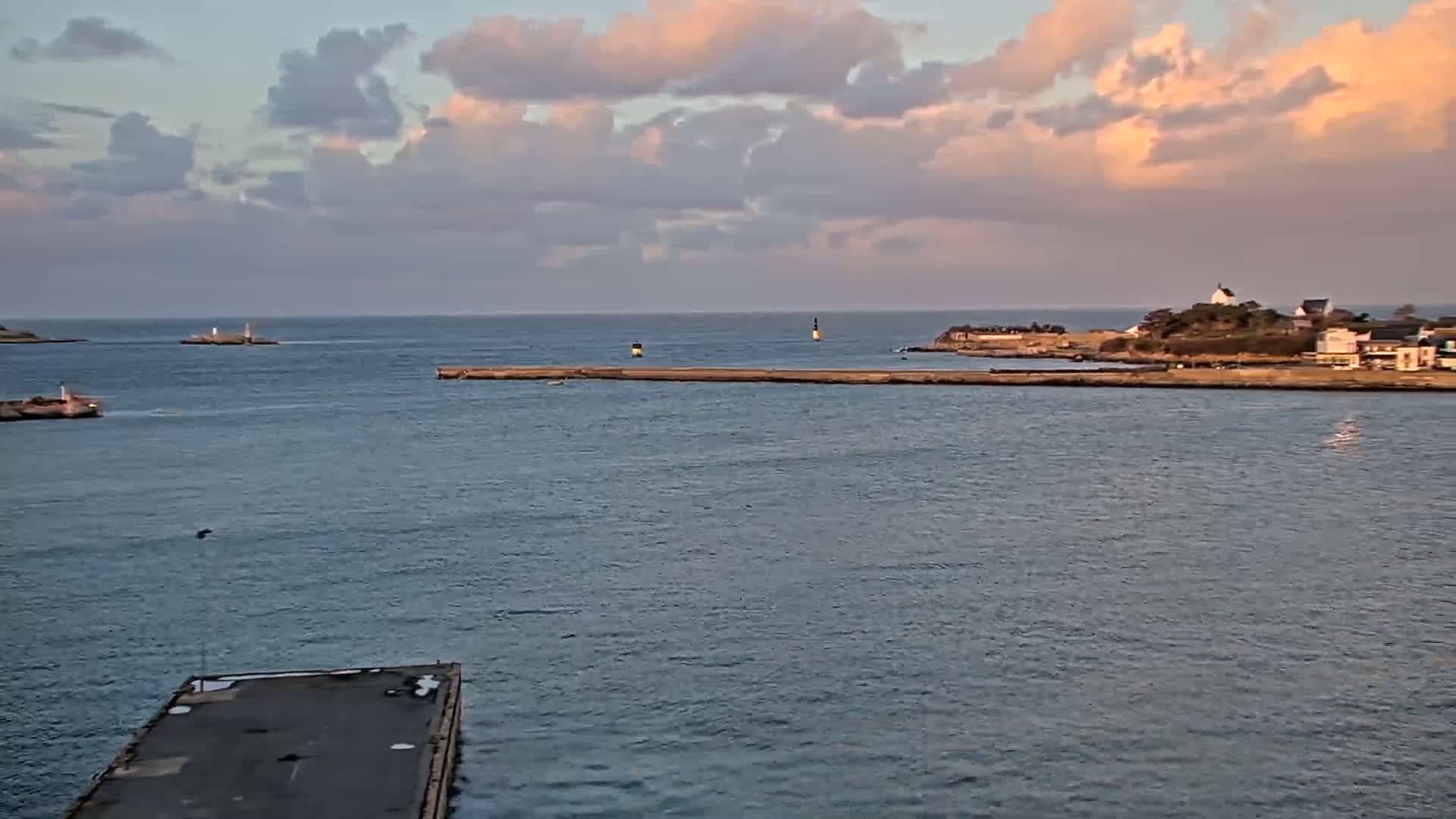 A harbor scene under a partly cloudy sky shows calm water, breakwaters, a small island with buildings, and a distant shoreline.