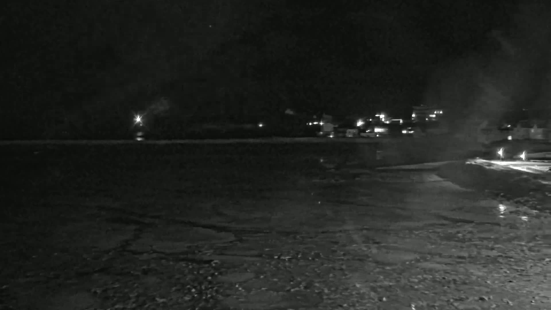 A nighttime view of a dark, icy body of water with a distant line of lights on the far shore under a dark, cloudy sky.