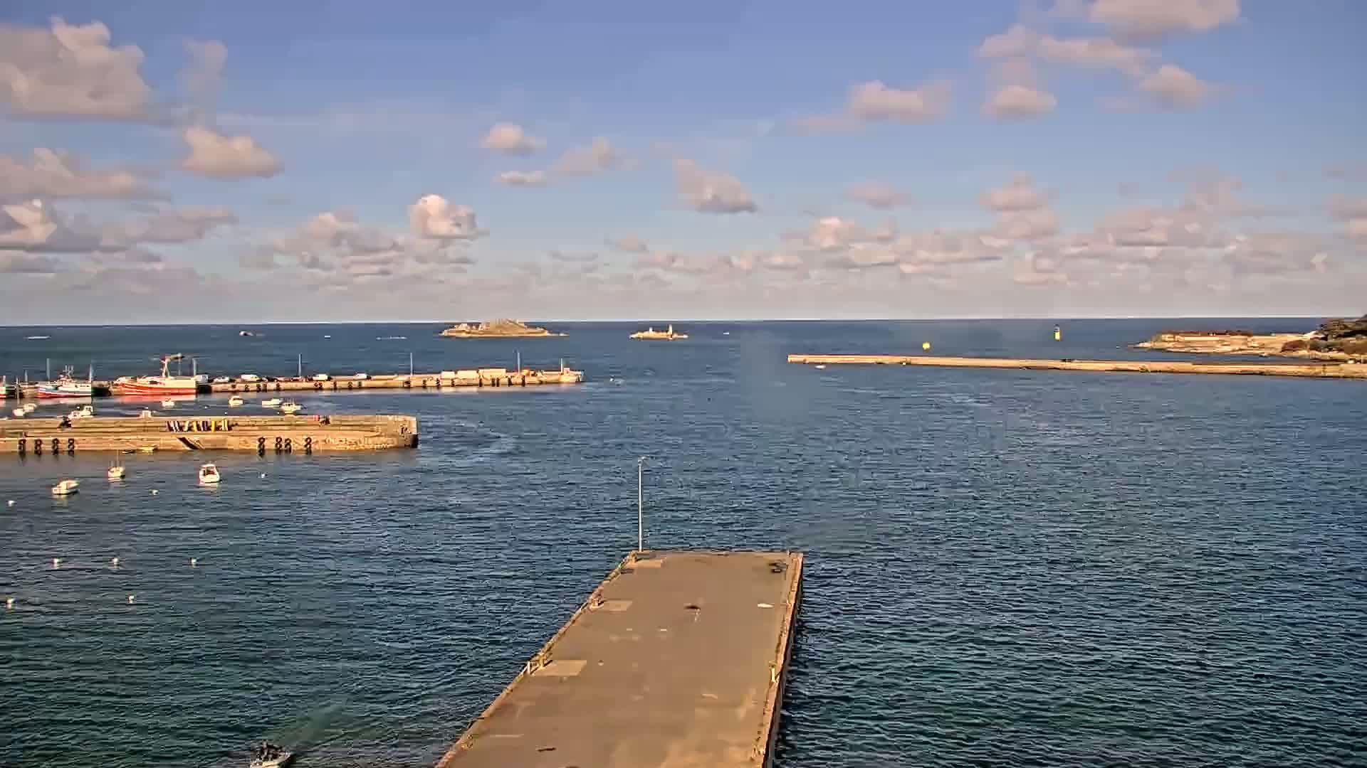 A harbor filled with boats is seen from above on a partly cloudy day, with a long pier extending into the water toward a small island in the distance.