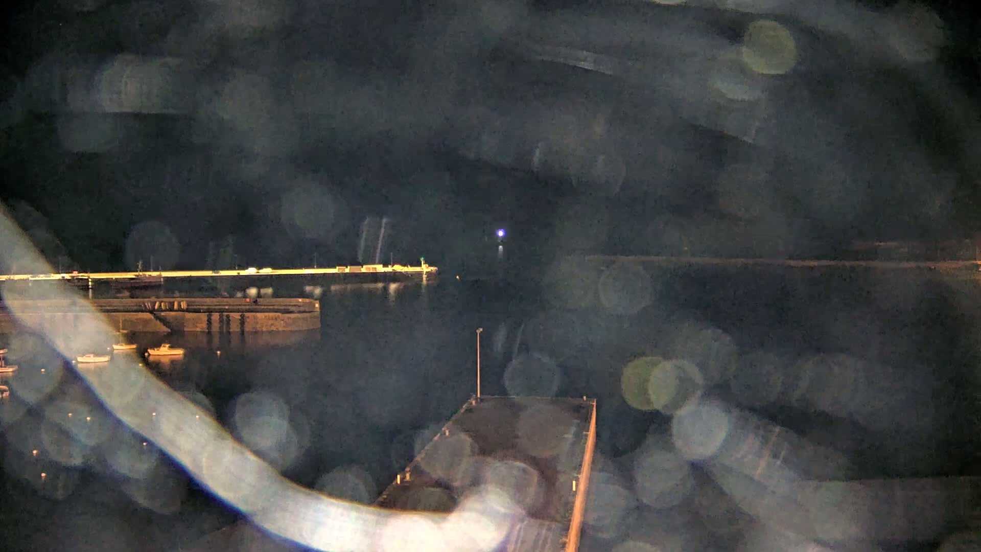 A nighttime view of a harbor with several small boats moored, a pier extending into the water, and lights visible in the distance, all seen through rain-streaked glass.
