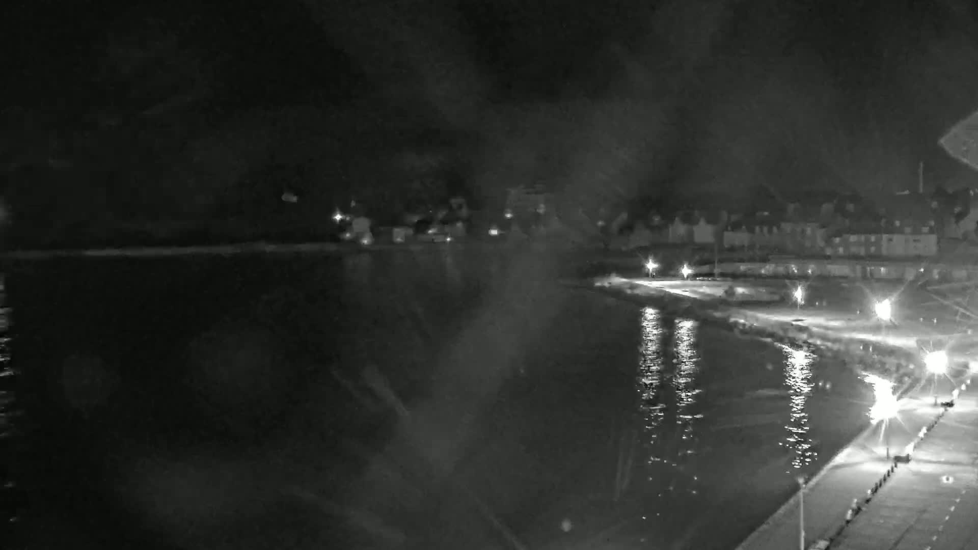 A nighttime view of a calm body of water next to a shoreline with buildings and streetlights visible in the distance.