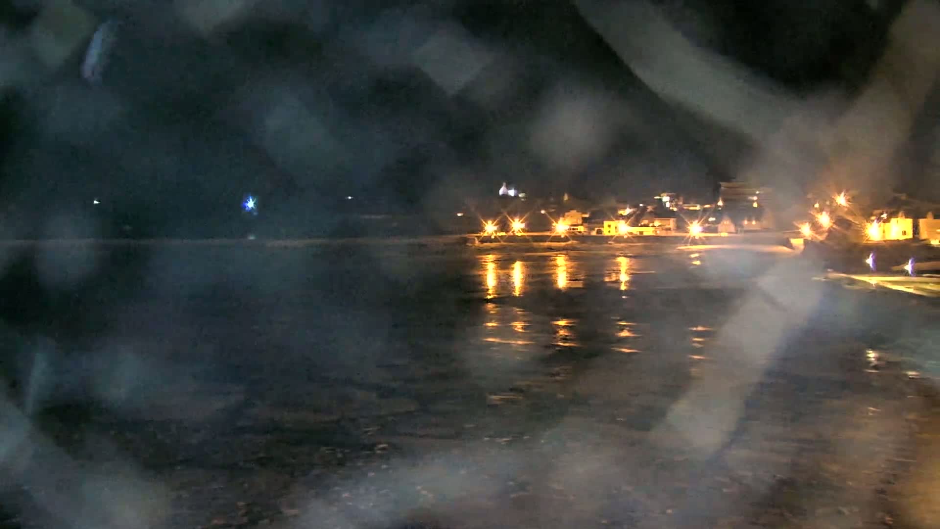 A nighttime view of a town's lights reflected on a body of water, seen through a rain-streaked window.