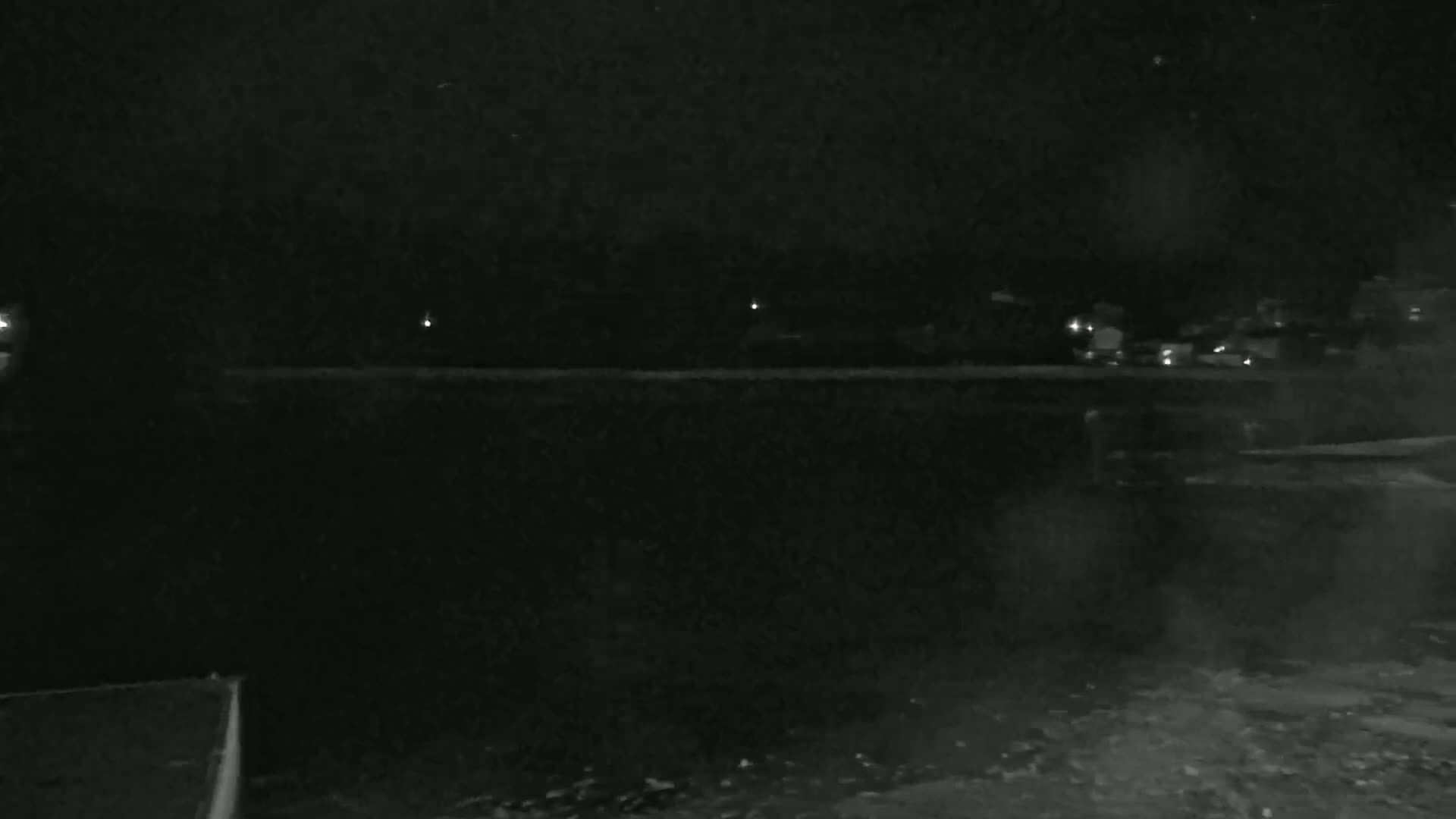 A dark, nighttime scene shows a body of water in the foreground, with a distant shoreline featuring a few faint lights under a cloudy sky.